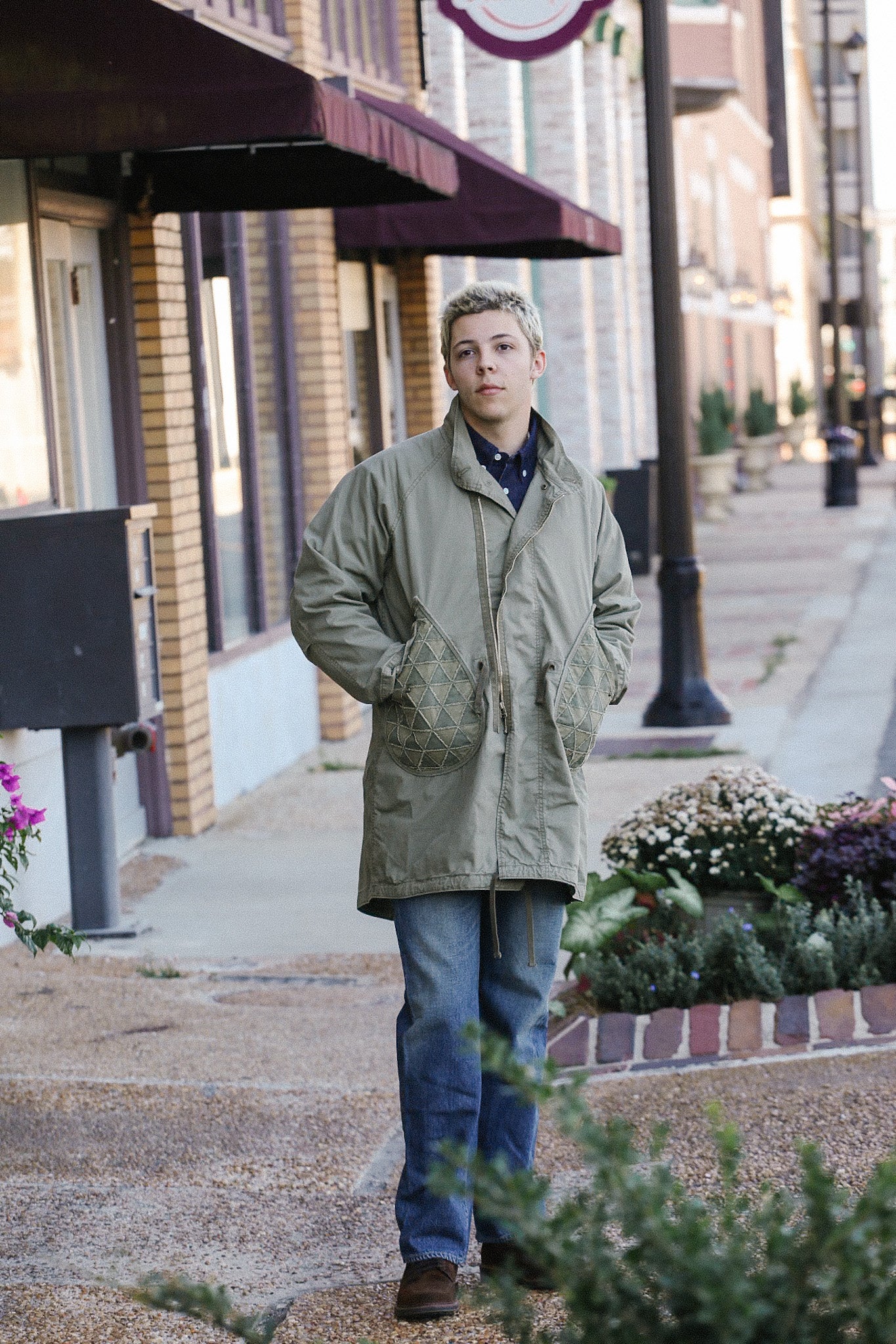 Young man wearing an olive green parka coat with quilted pockets walking on a city sidewalk