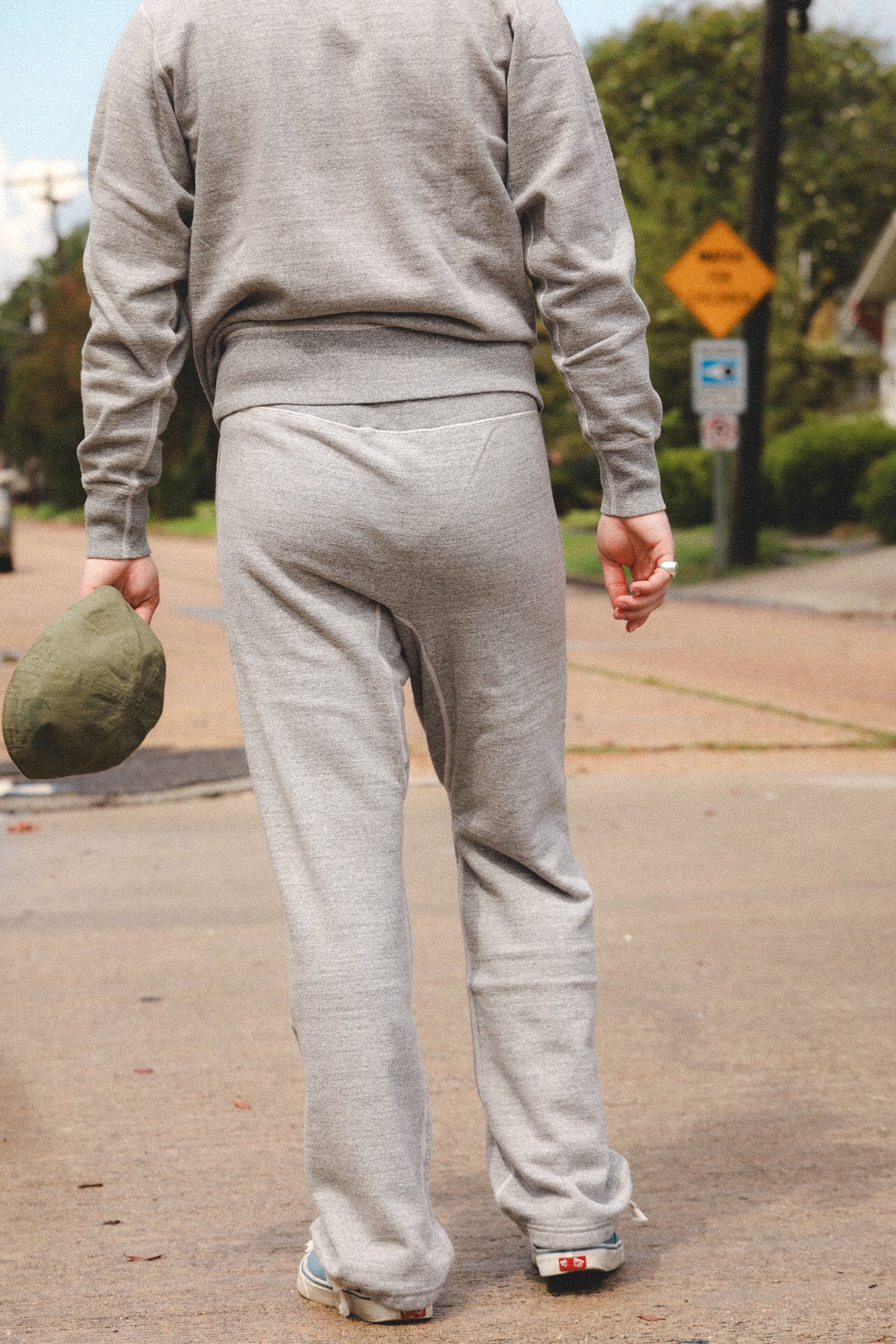 A person viewed from behind wearing a light gray heather sweatshirt and matching sweatpants, walking outdoors on a paved street with trees and a sign in the background.