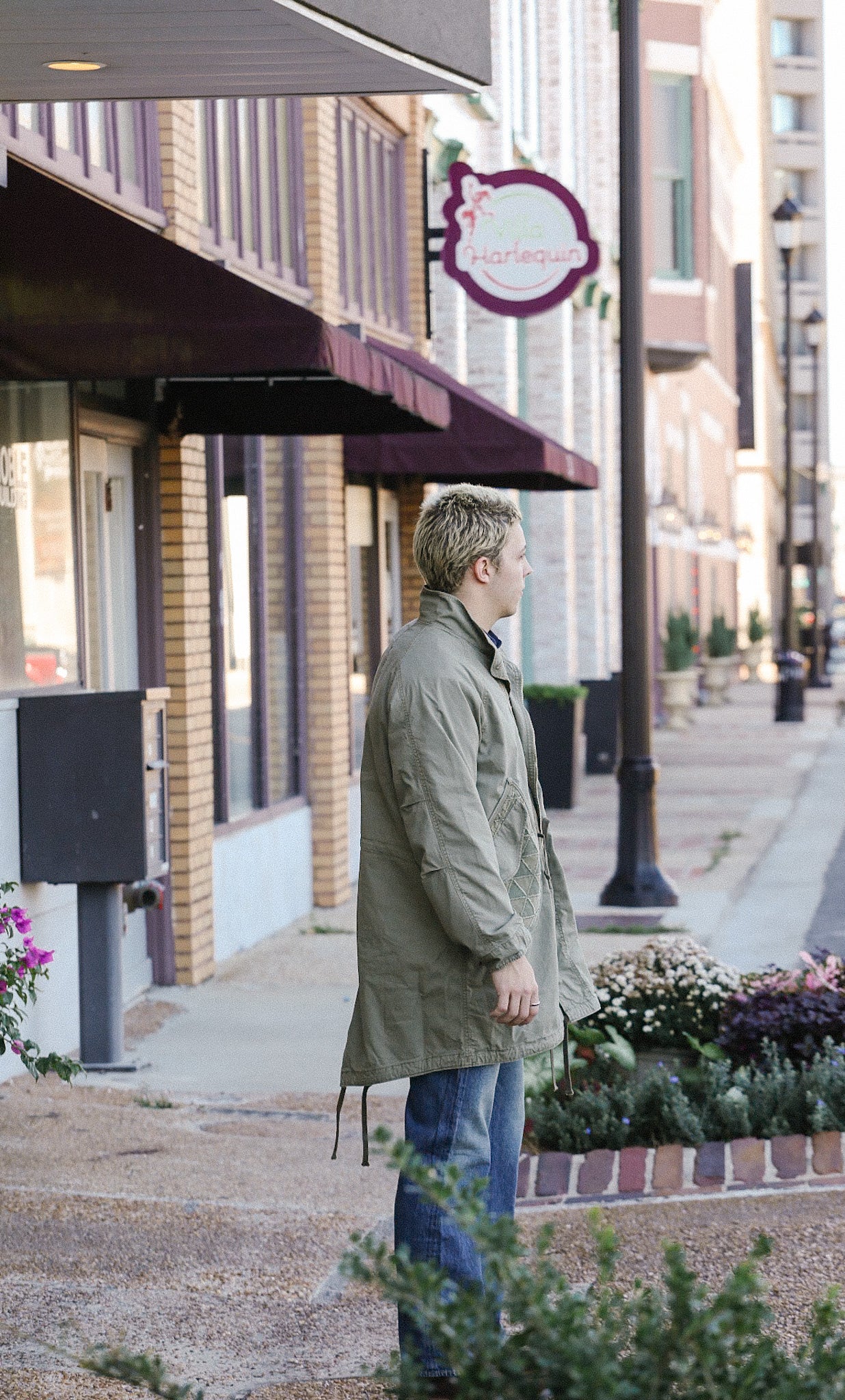 Man wearing olive green jacket and jeans standing on downtown sidewalk near shops and flower beds