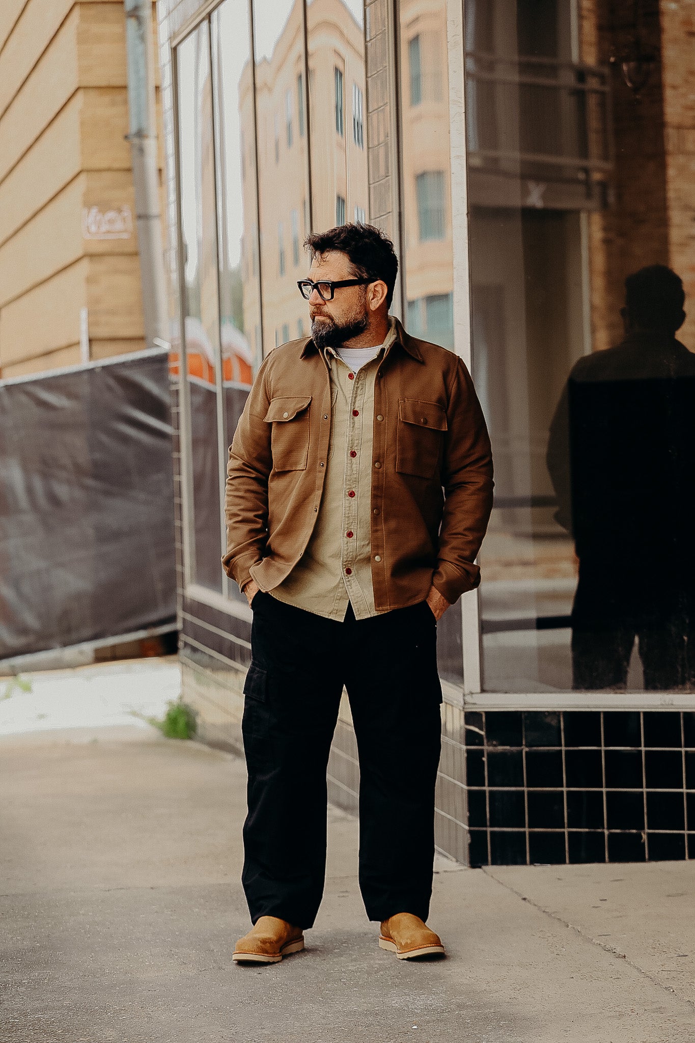 Man wearing brown jacket, beige shirt, black pants, and tan boots standing on urban sidewalk