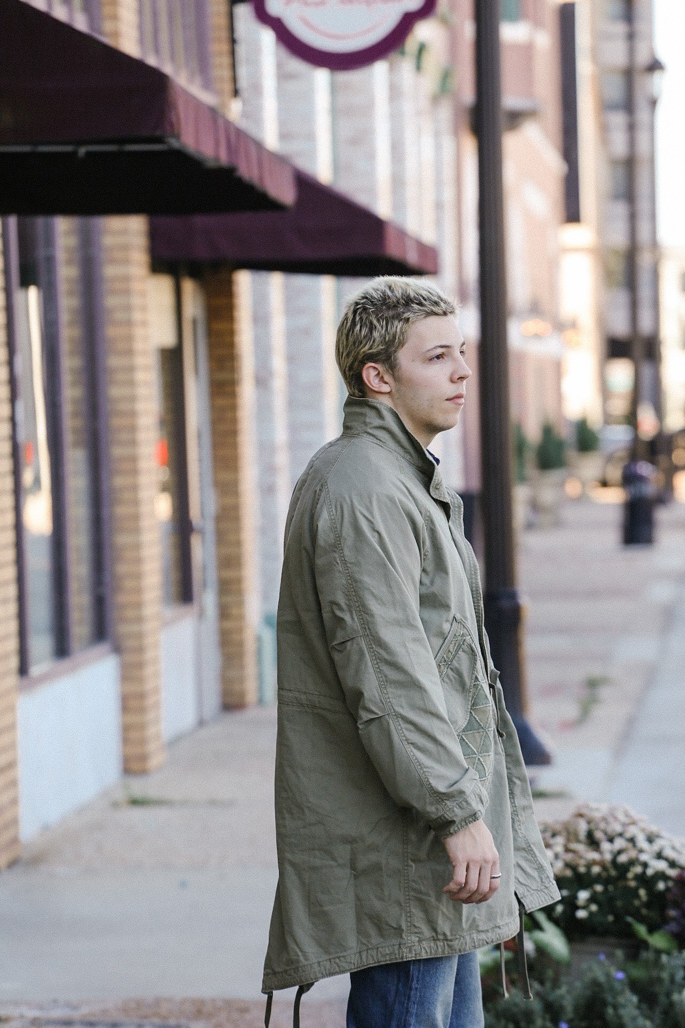 Young man wearing olive green military-style jacket standing on urban sidewalk near brick buildings