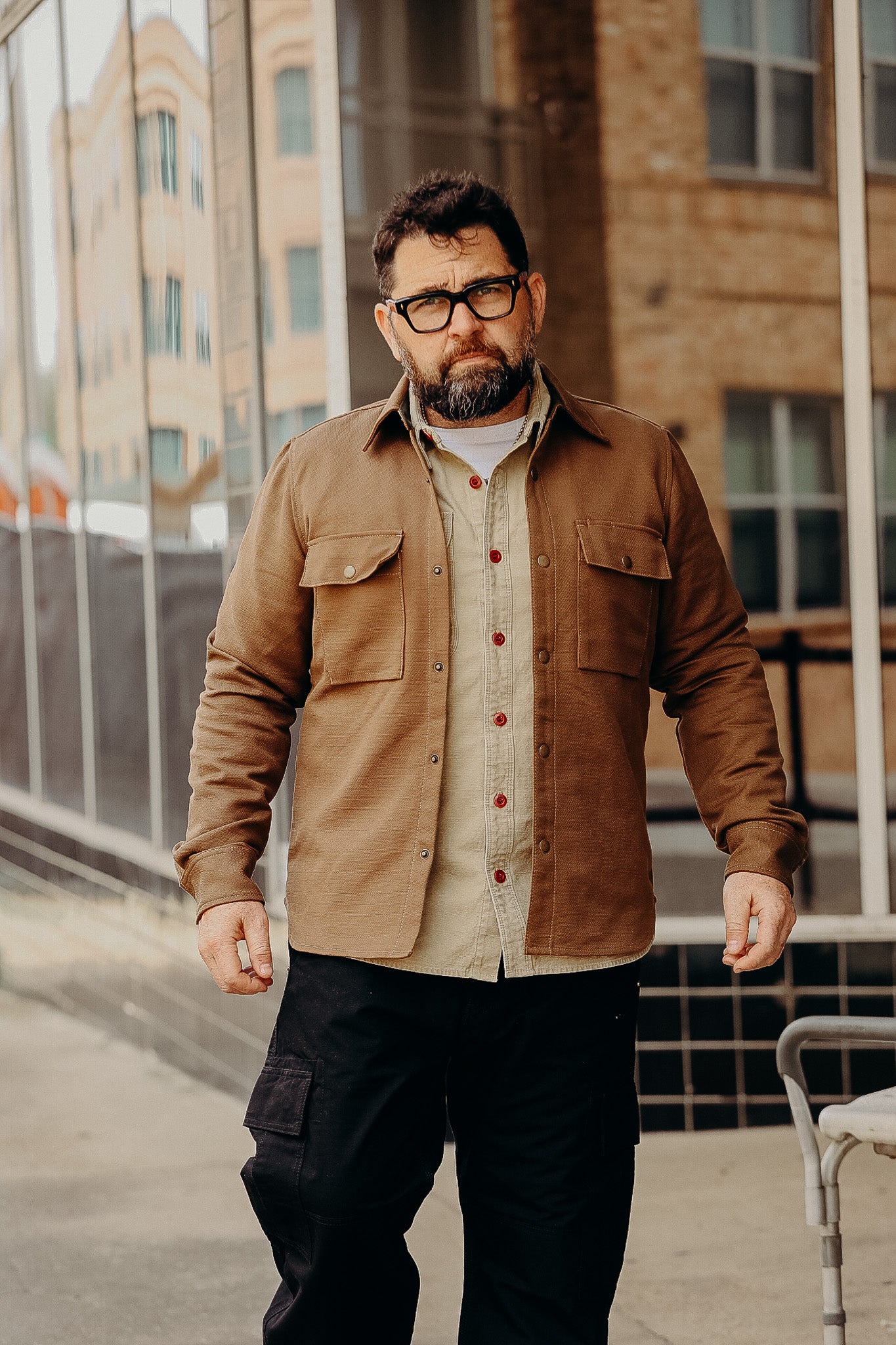 Bearded man wearing brown work jacket and beige shirt standing in urban outdoor setting