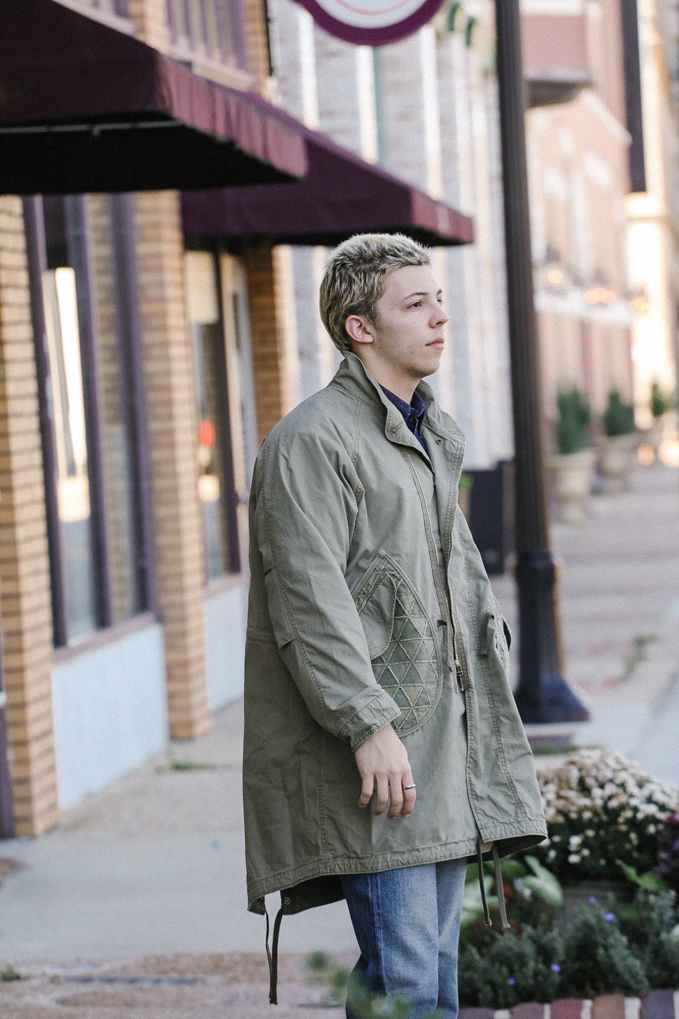 Young man wearing olive green military-style jacket standing on urban sidewalk with brick buildings