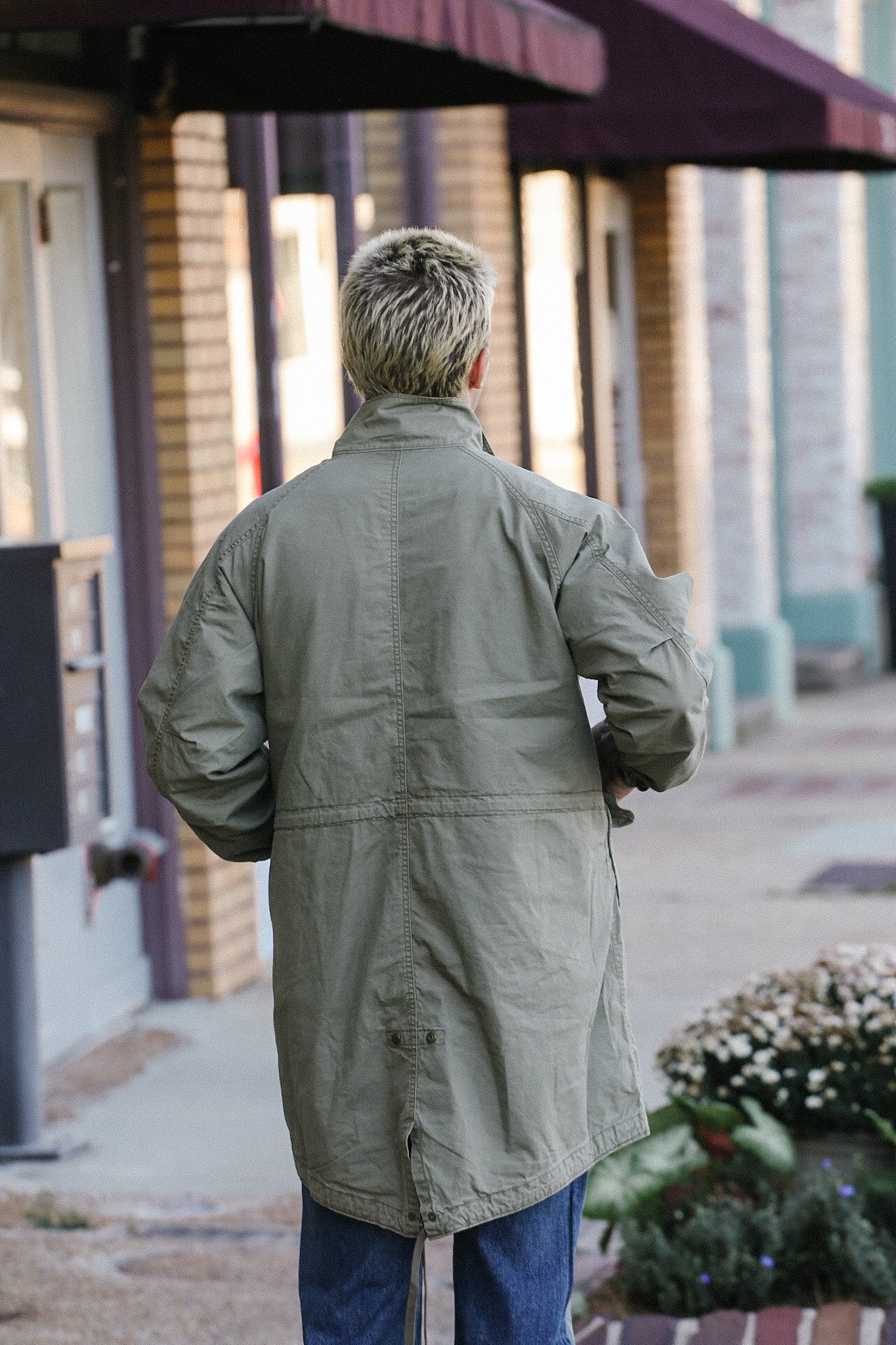 Back view of person wearing long olive green jacket and blue jeans walking in urban street