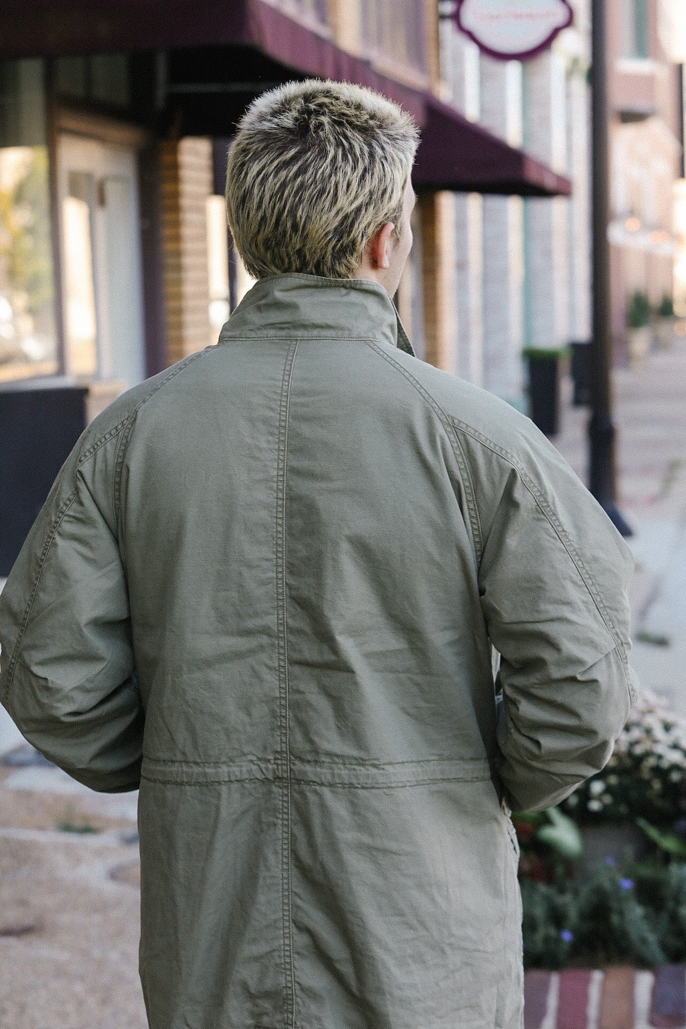 Back view of person wearing olive green casual jacket walking on urban sidewalk near storefronts