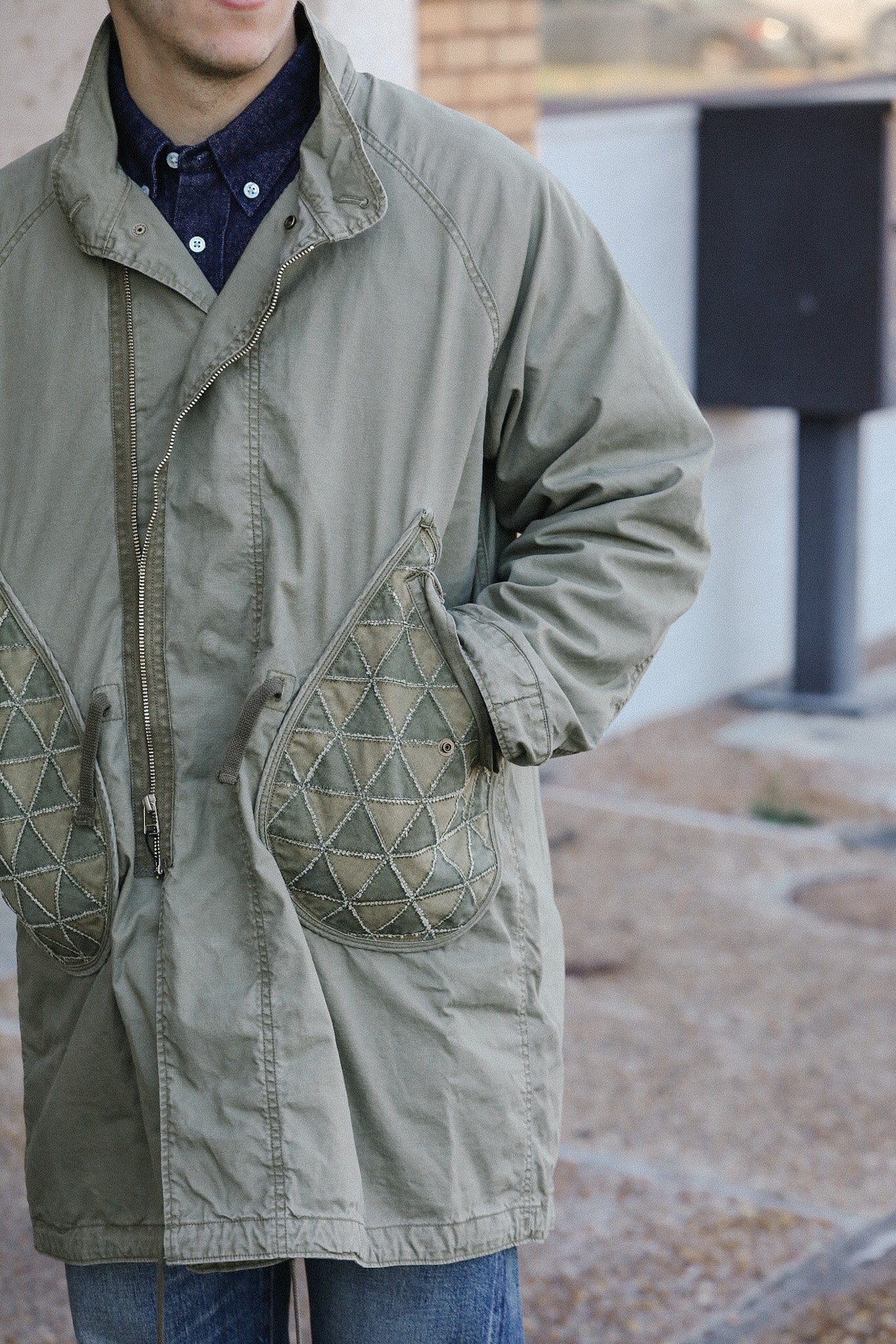 Close-up of a person wearing a vintage olive green jacket with quilted patch pockets and blue jeans outdoors