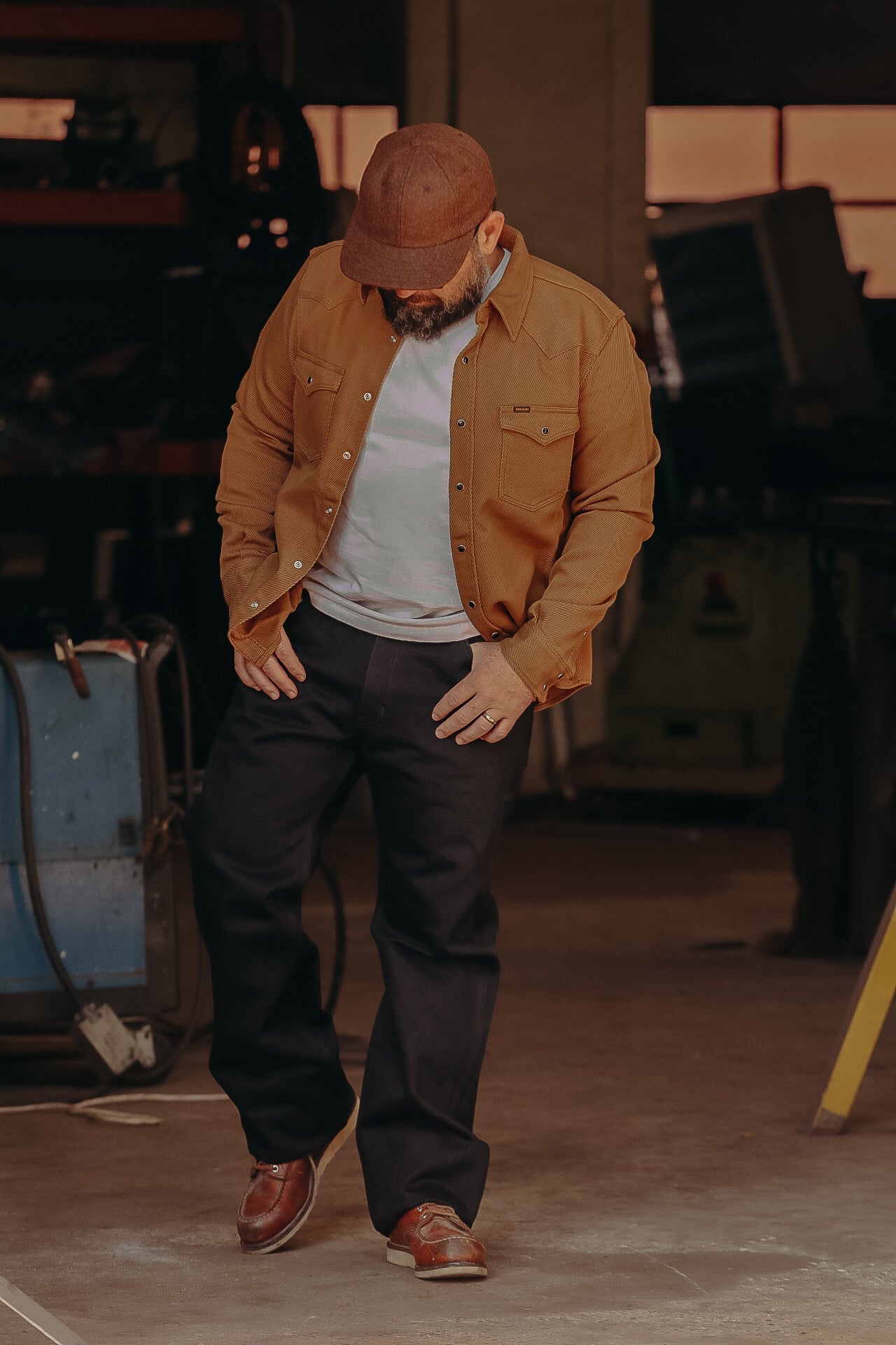 Man in brown cap, rugged brown jacket, white t-shirt, black pants, and brown shoes in industrial workshop