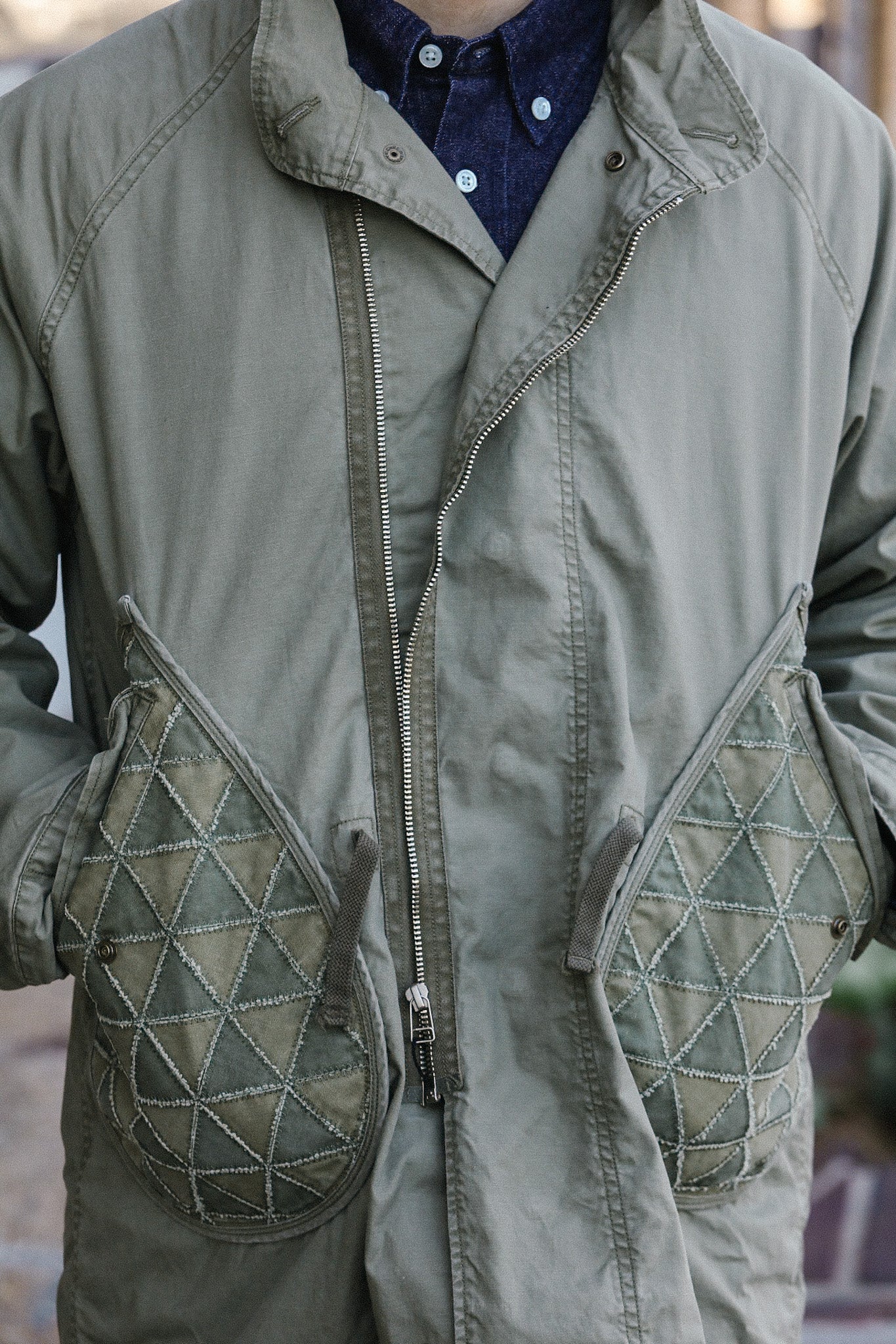 Close-up of gray-green jacket with quilted diamond pattern pockets and zipper, layered over blue button-up shirt