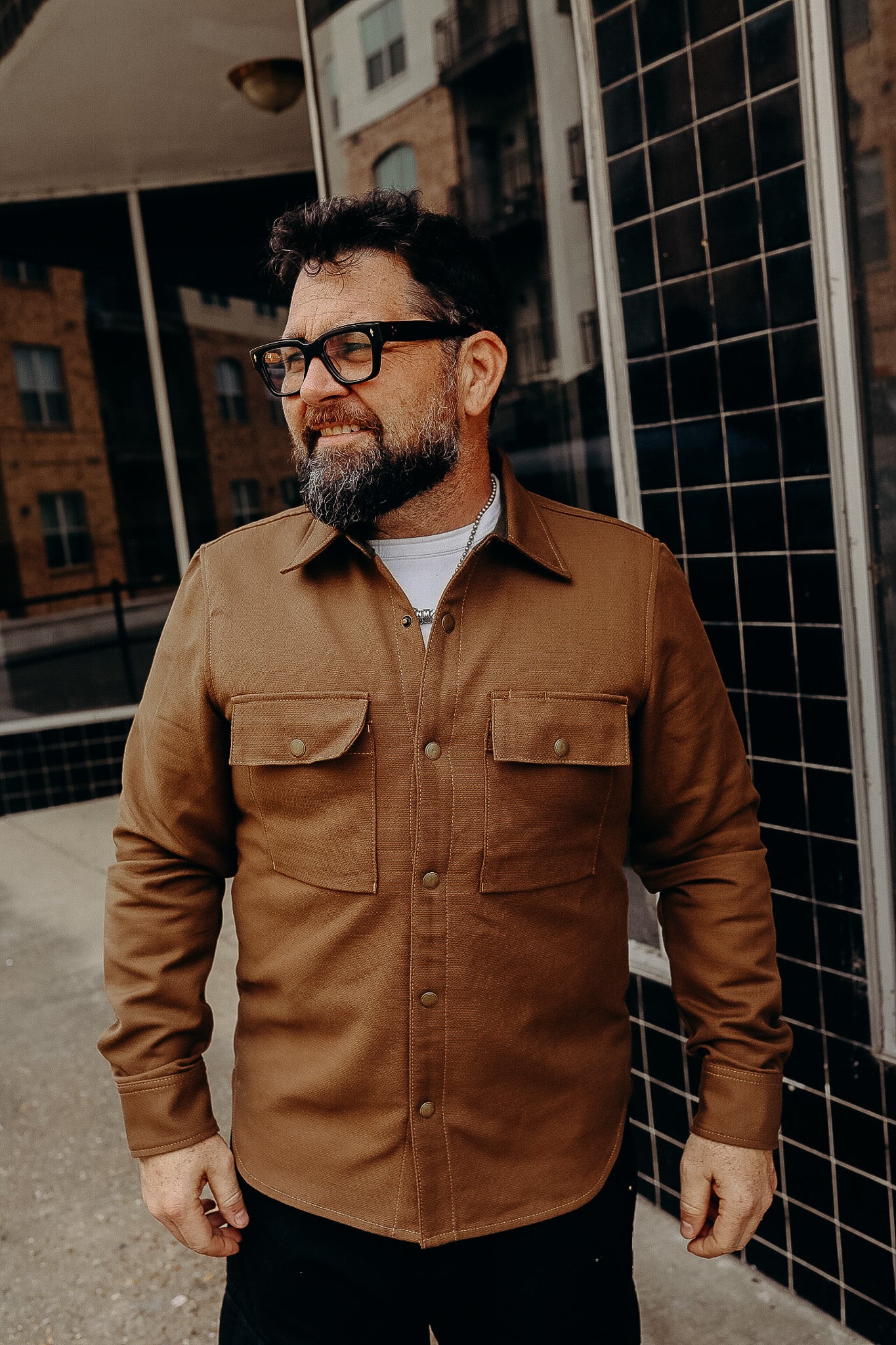 Bearded man wearing brown work shirt with snap pockets standing outdoors near building with black tiled wall