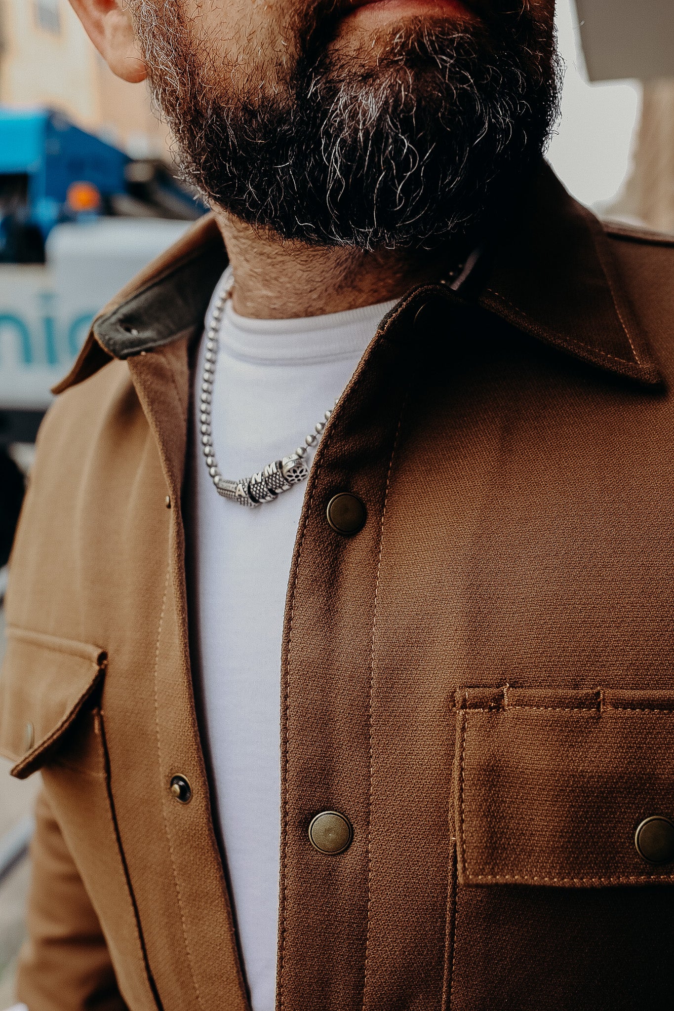 Close-up of a man with a dark beard wearing a brown button-up jacket and a silver beaded necklace over a white shirt