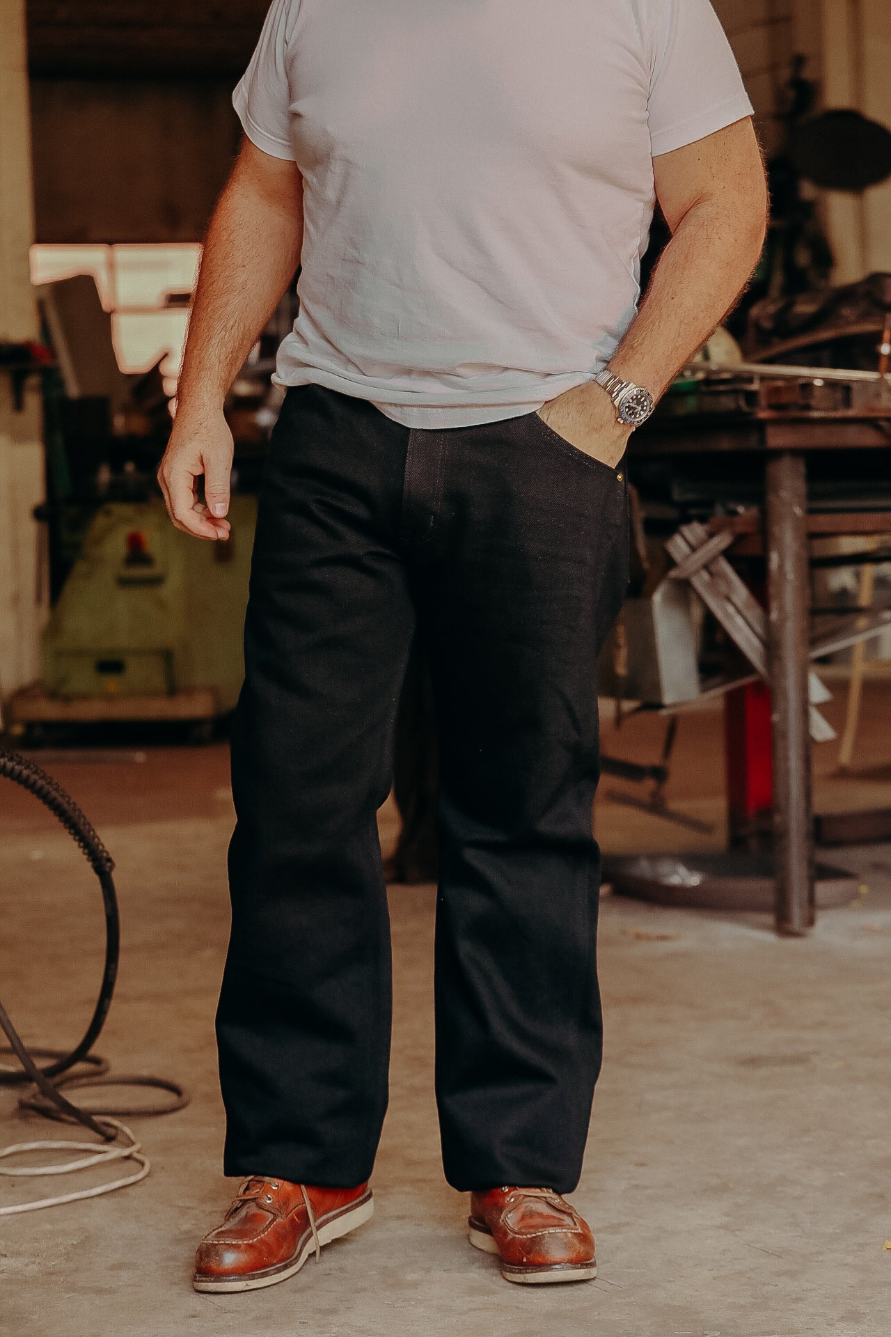 Man wearing black work pants, white t-shirt, and brown leather boots in a workshop setting