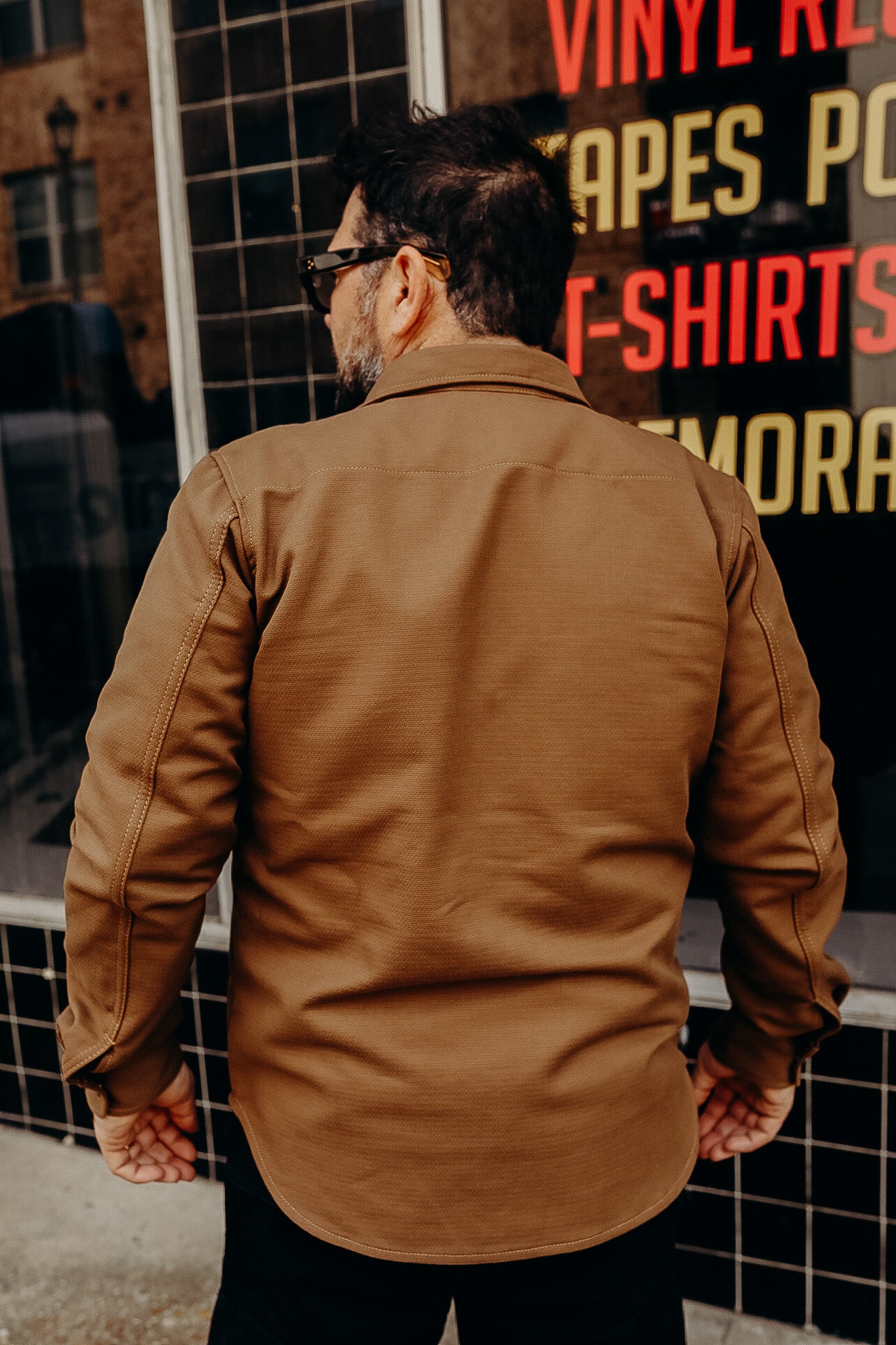 Man wearing a brown rugged jacket standing outdoors near a tiled wall and a store window with colorful text