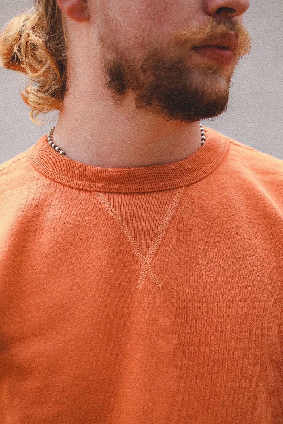 Close-up of a man wearing a textured orange crewneck sweatshirt, showing the chest area with a subtle tonal embroidered V-neck design.