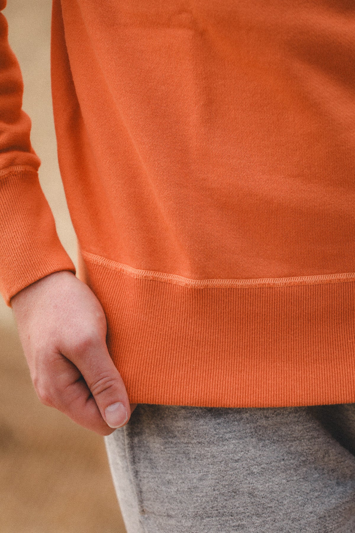 Close-up of a person wearing a bright orange ribbed sweatshirt, showing the hand and part of the gray sweatpants against a softly blurred tan background.