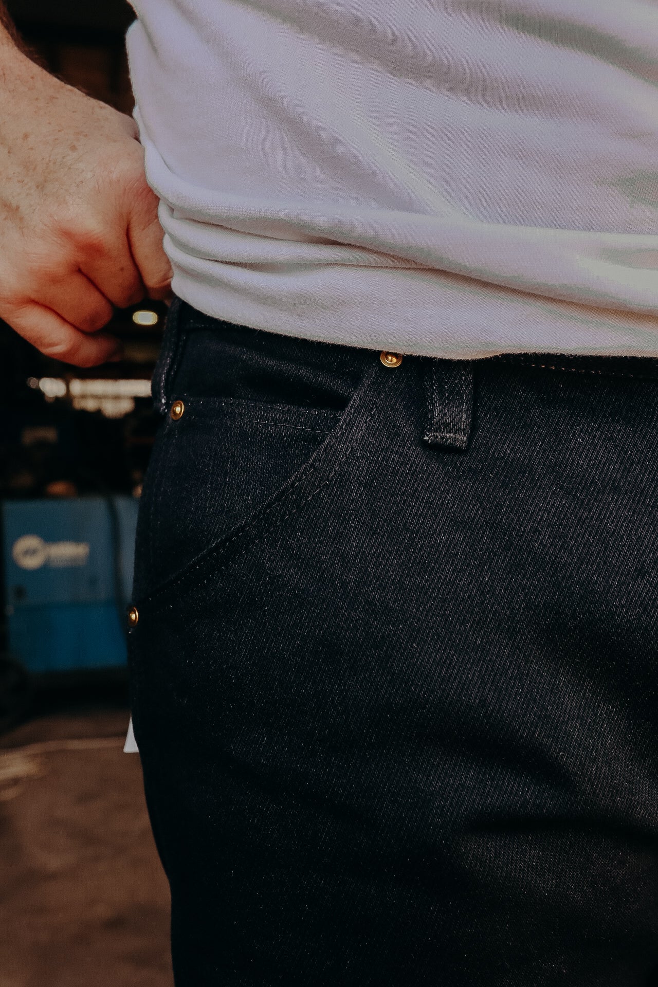 Close-up of person wearing dark denim jeans with brass rivets and a white T-shirt in workshop setting