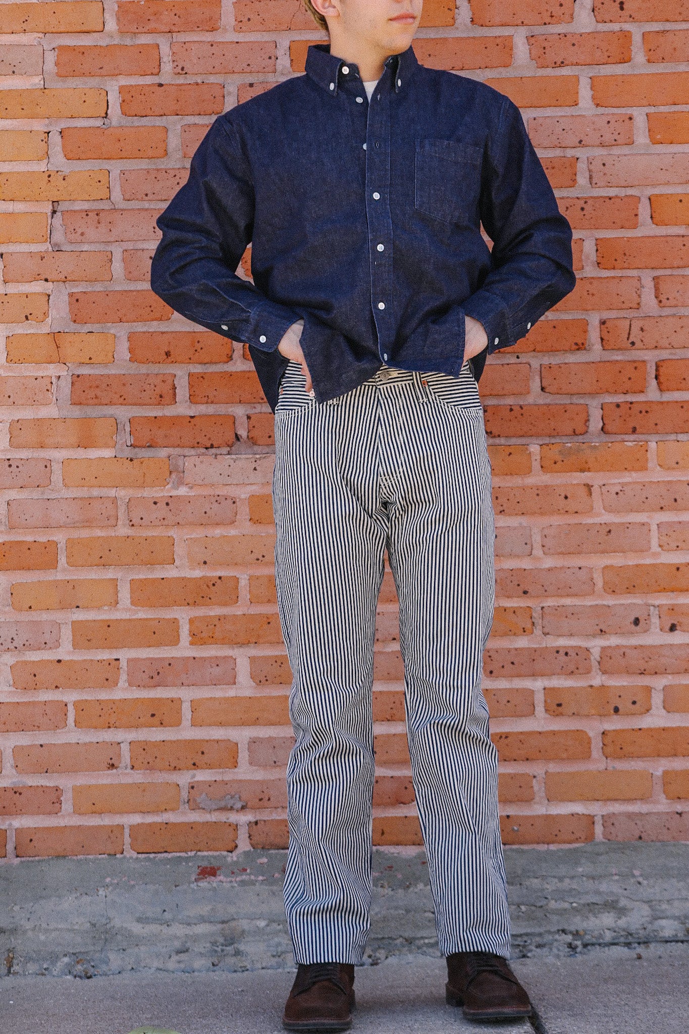 Man wearing navy button-up shirt and black-and-white striped pants standing against brick wall