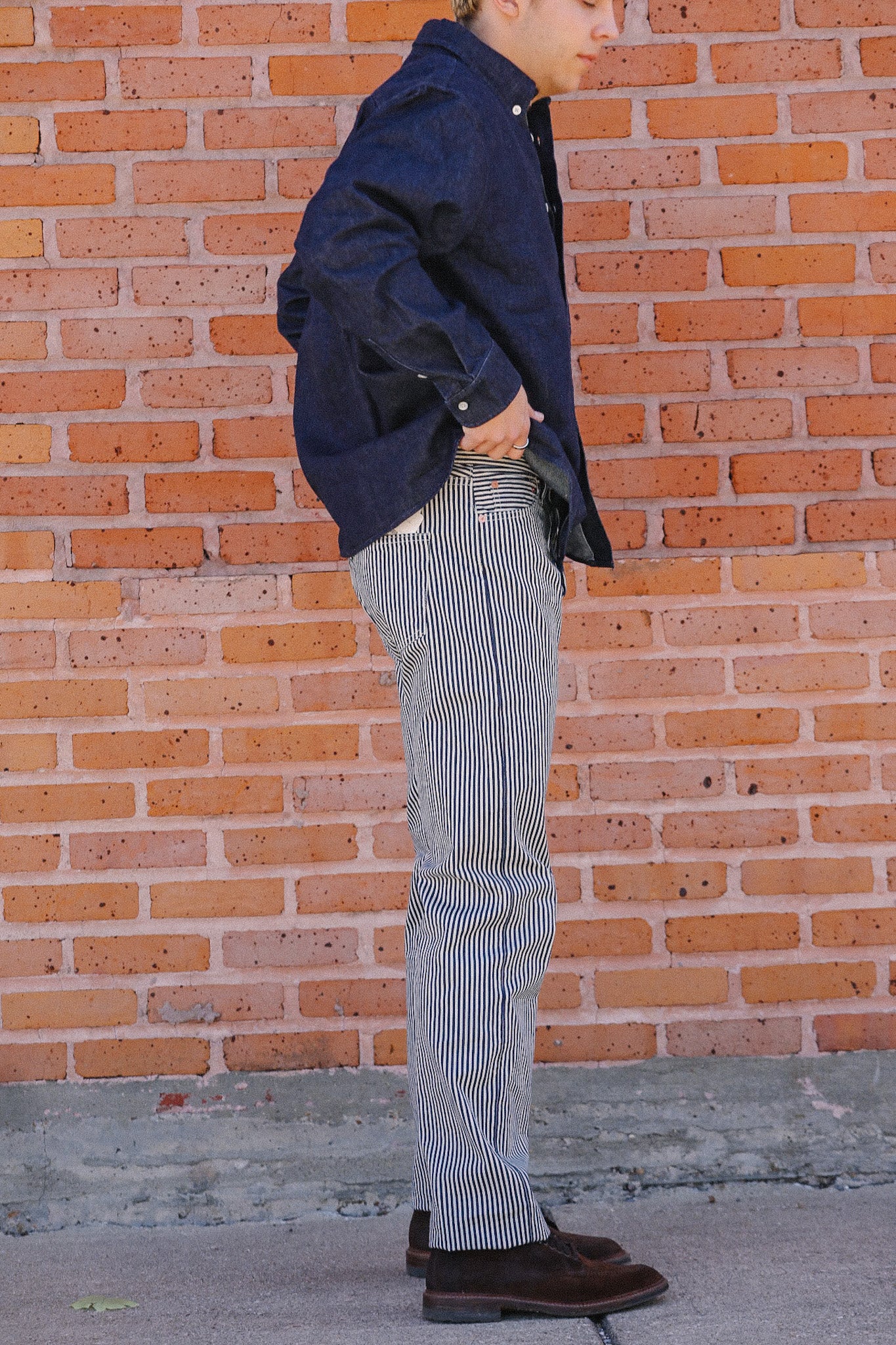 Man in navy blue shirt and striped denim jeans standing against a brick wall with brown leather shoes