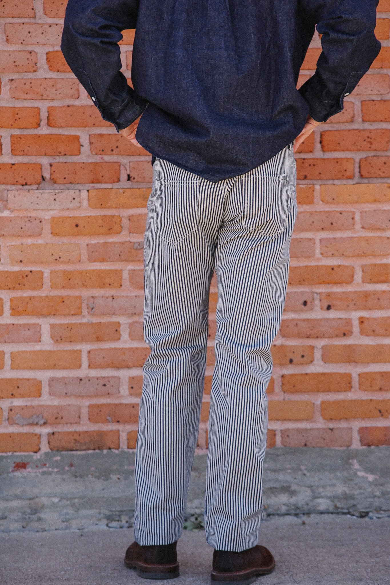 Person wearing black and white striped pants and dark denim shirt standing against a brick wall