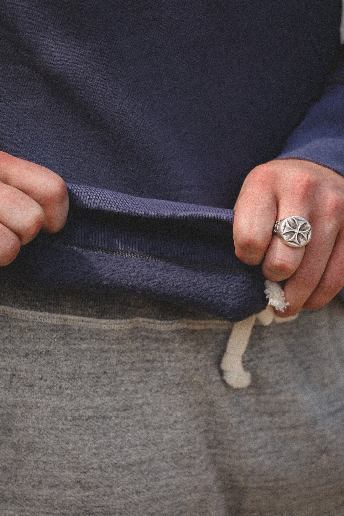 Close-up of a person wearing a navy blue sweatshirt and gray sweatpants, pulling the sweatshirt cuff down slightly to show the fabric and white drawstring. A silver ring with a geometric star design is worn on the ring finger.