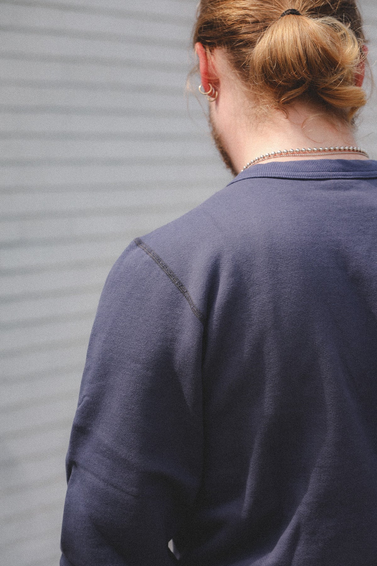 A person viewed from behind wearing a dark slate blue crewneck sweatshirt, standing against a gray corrugated metal wall.