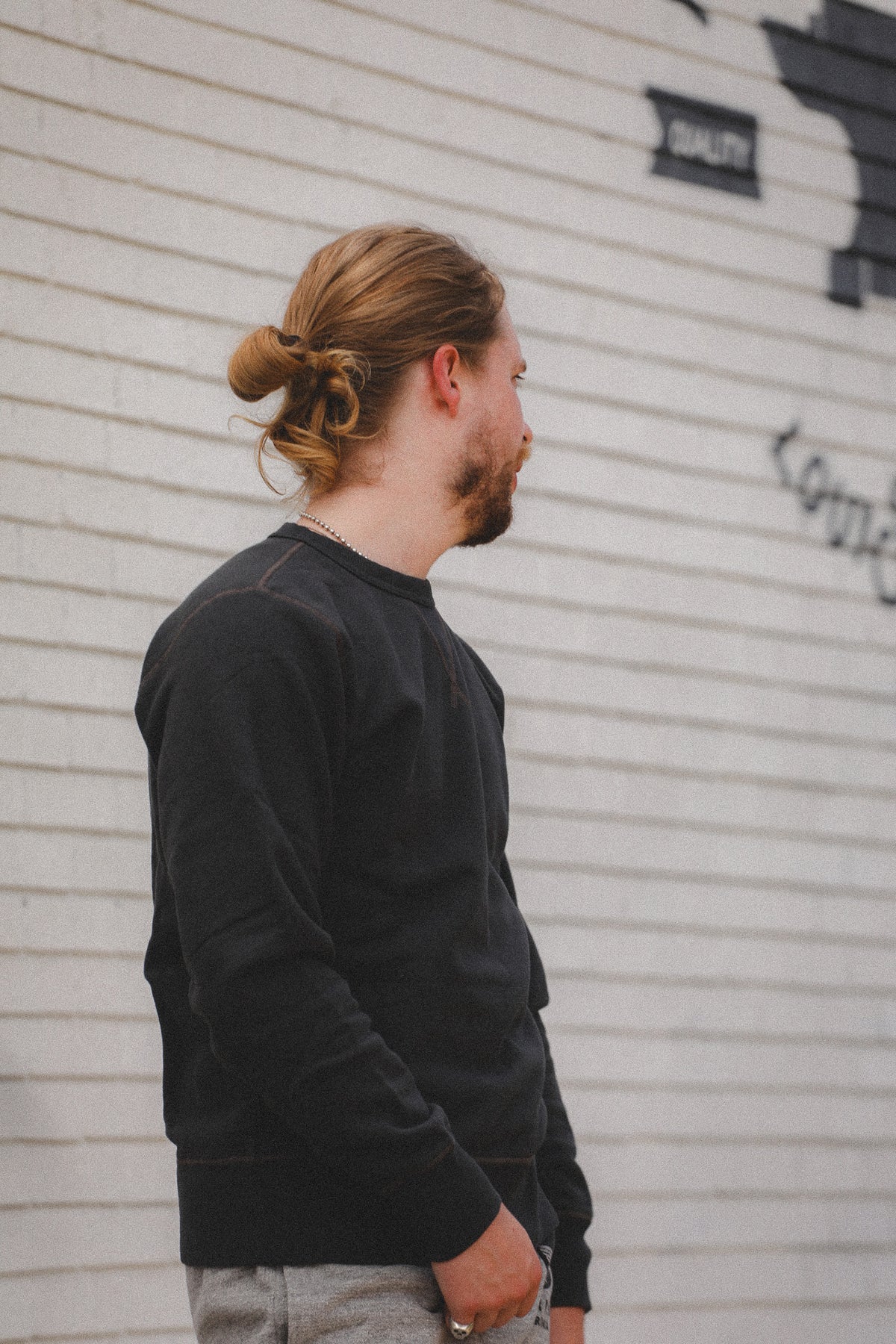 A man with a bun hairstyle and beard, wearing a black crewneck sweatshirt and gray pants, standing in profile against a white brick wall.