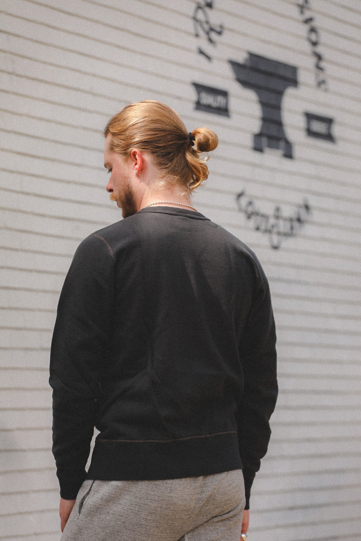 A man with blonde hair in a bun is shown from behind wearing a black crewneck sweatshirt and gray sweatpants, standing in front of a white corrugated metal wall with black graffiti.