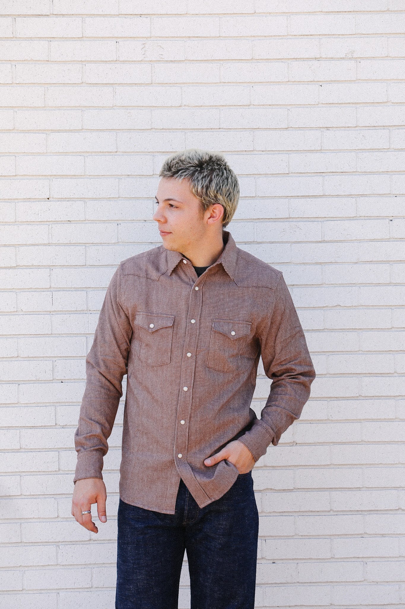 Man wearing brown corduroy shirt and dark jeans standing against white brick wall