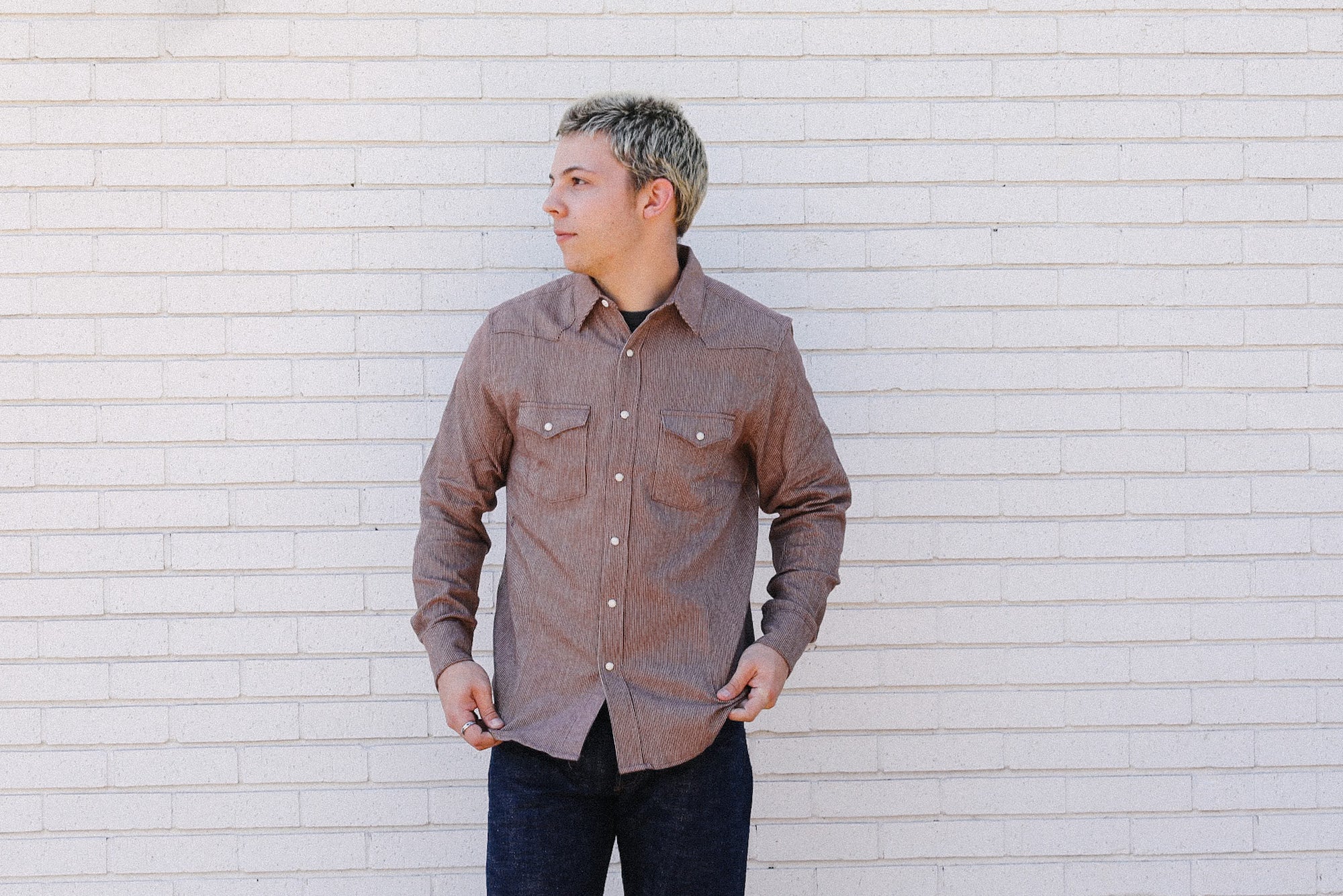 Man wearing brown button-up shirt and dark jeans standing against white brick wall