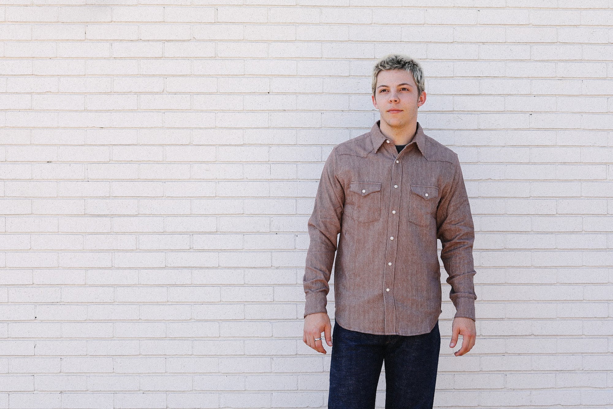Man wearing brown long-sleeve button-up shirt and dark jeans standing against white brick wall