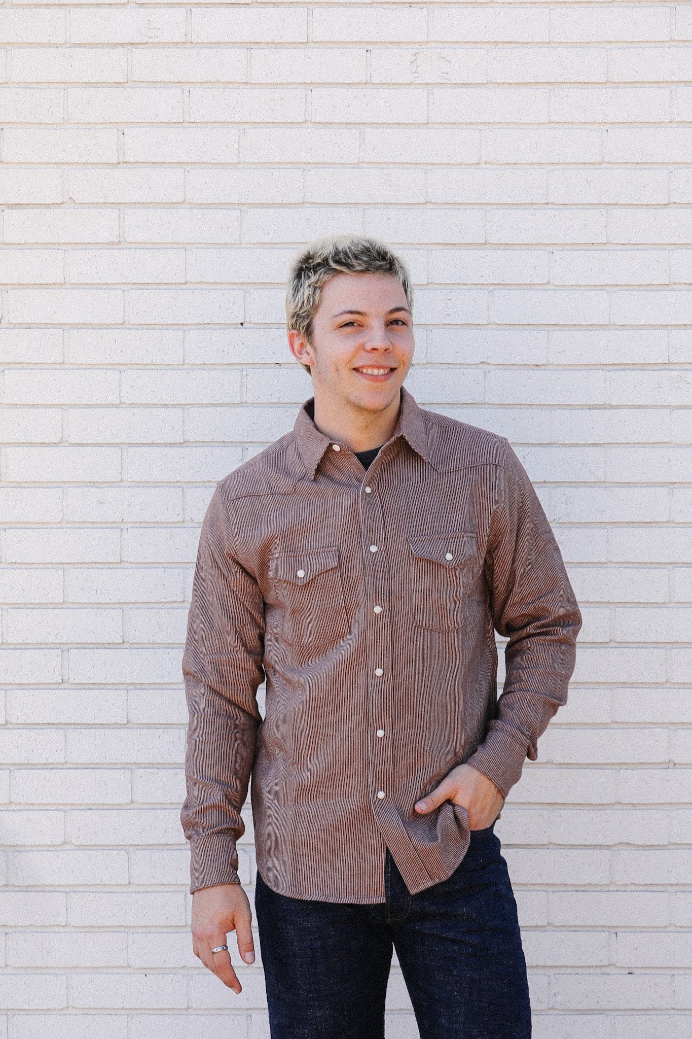 Man wearing brown corduroy button-up shirt and dark jeans standing against white brick wall