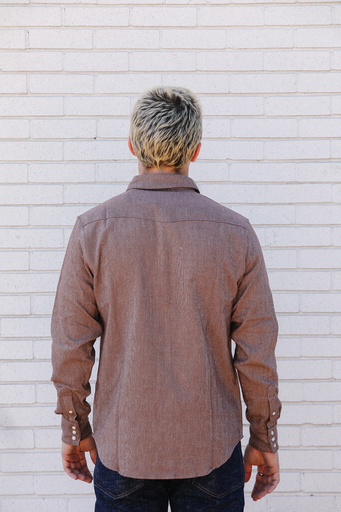 Man wearing brown striped work shirt standing against a white brick wall, back view
