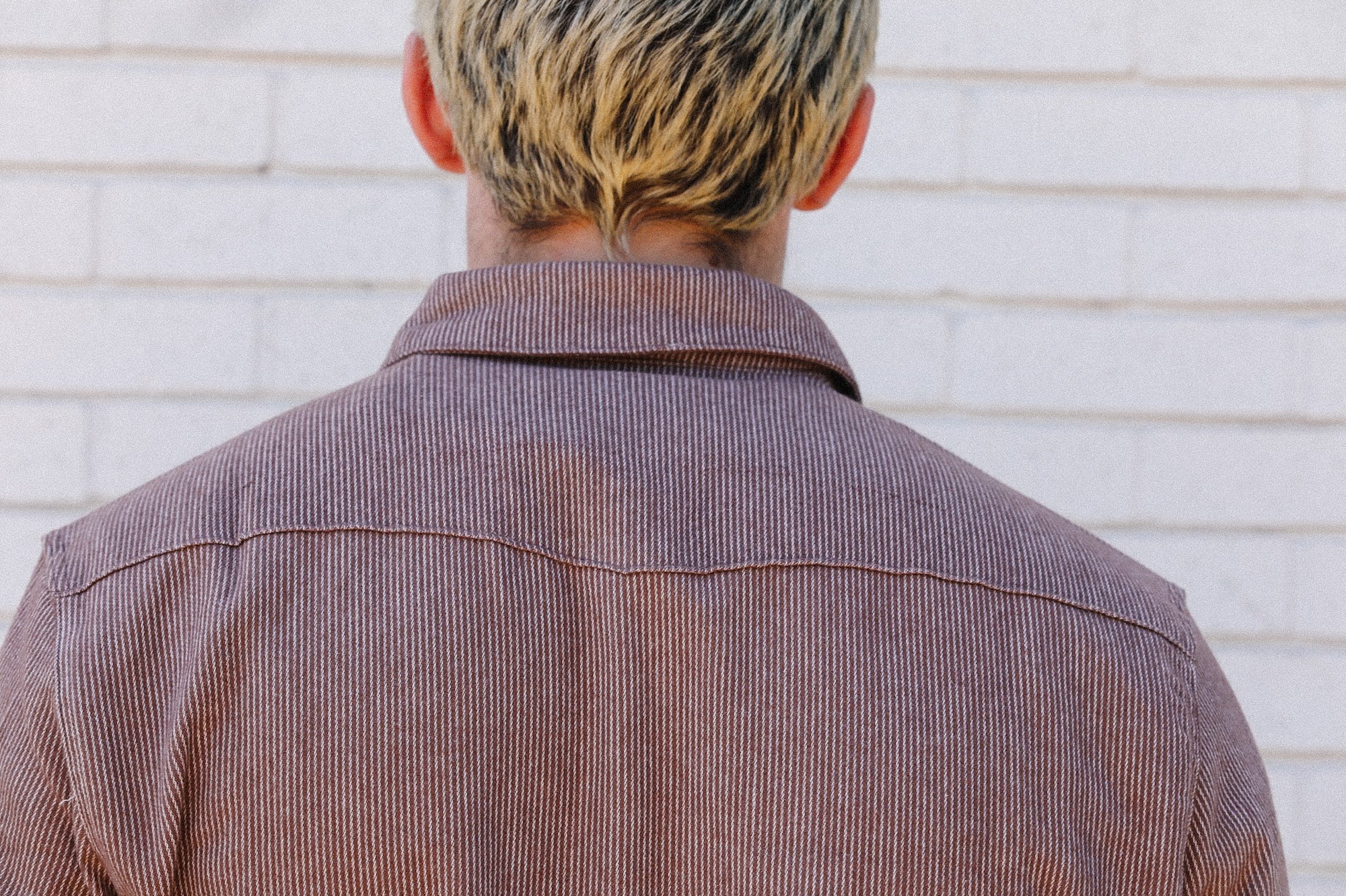 Back view of person with short blond hair wearing a brown striped jacket against white brick wall