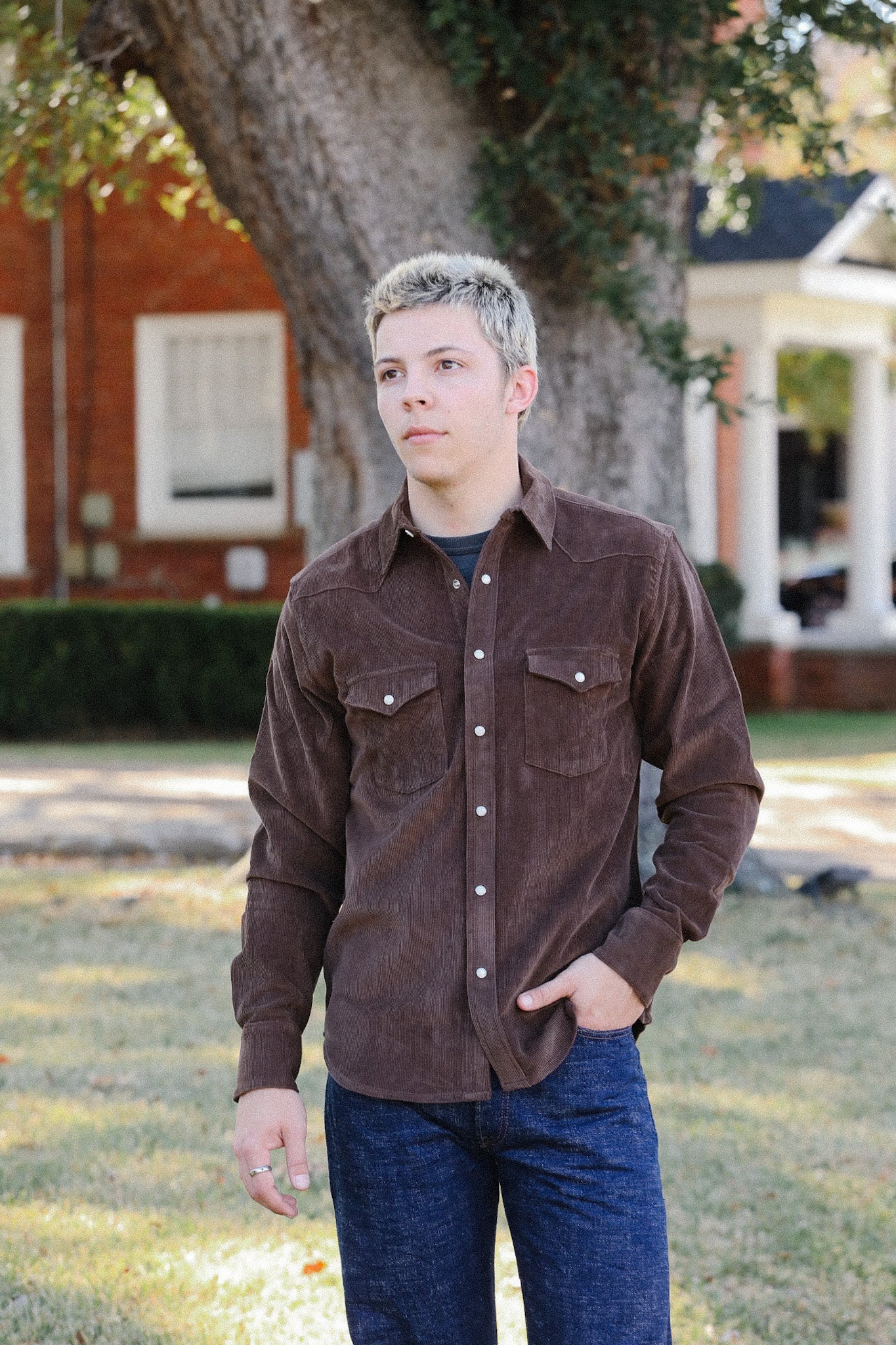 Young man wearing brown corduroy button-up shirt and blue jeans standing outdoors by large tree
