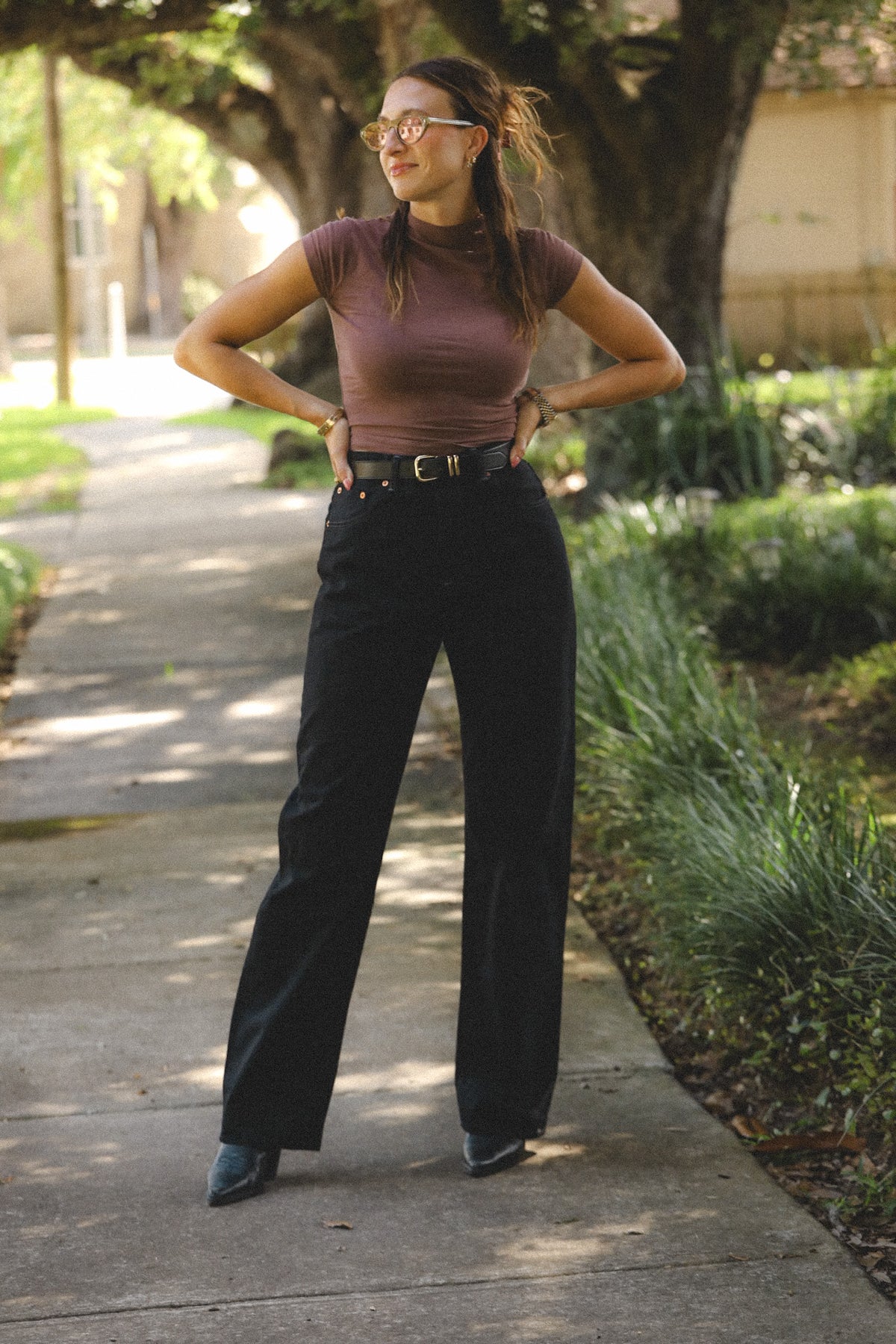 A woman stands on a sunlit sidewalk outdoors, wearing a fitted mauve-brown short-sleeve mock-neck top, black wide-leg pants with a black belt, and dark pointed shoes.