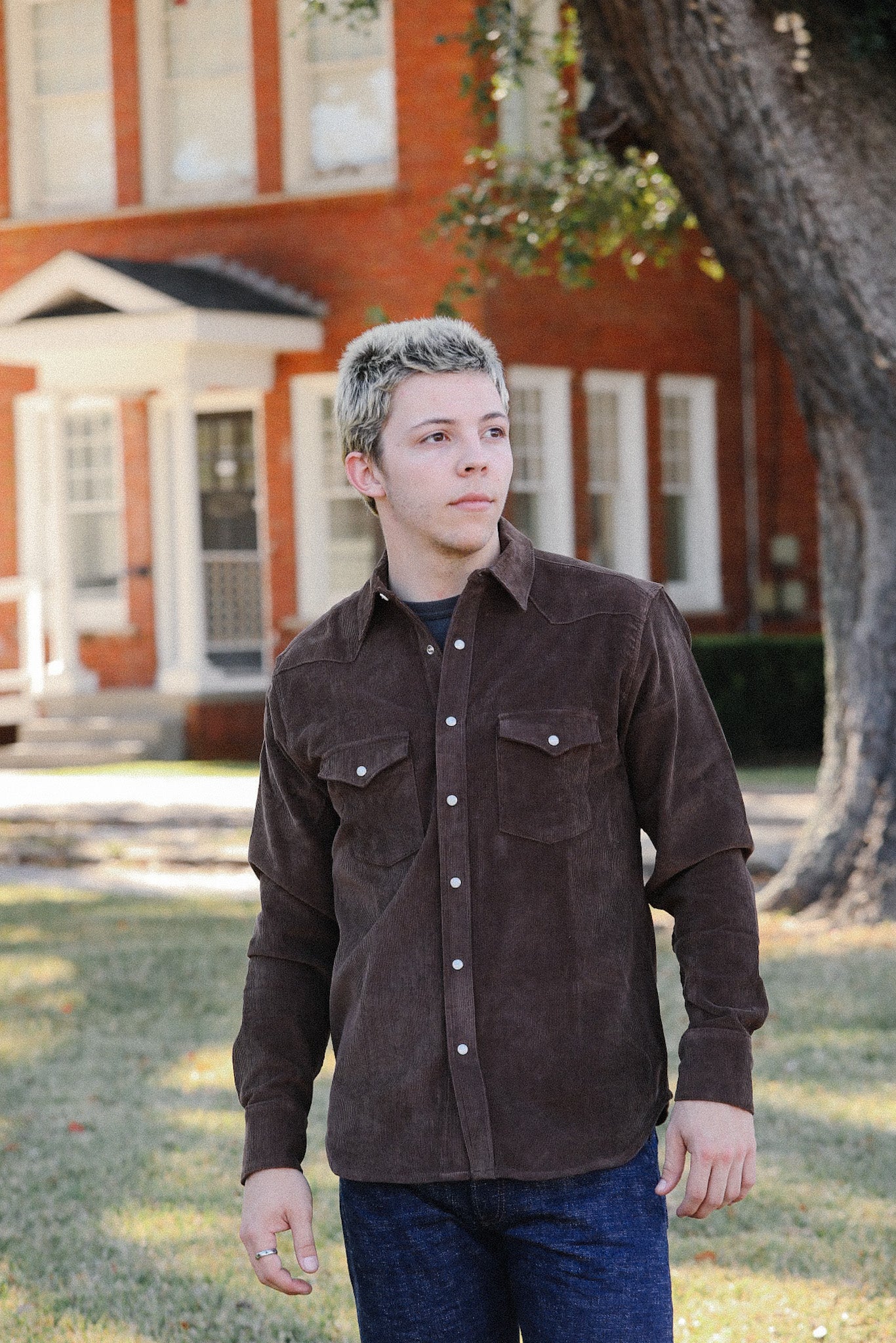 Young man wearing brown corduroy shirt and jeans standing outdoors near tree and brick building