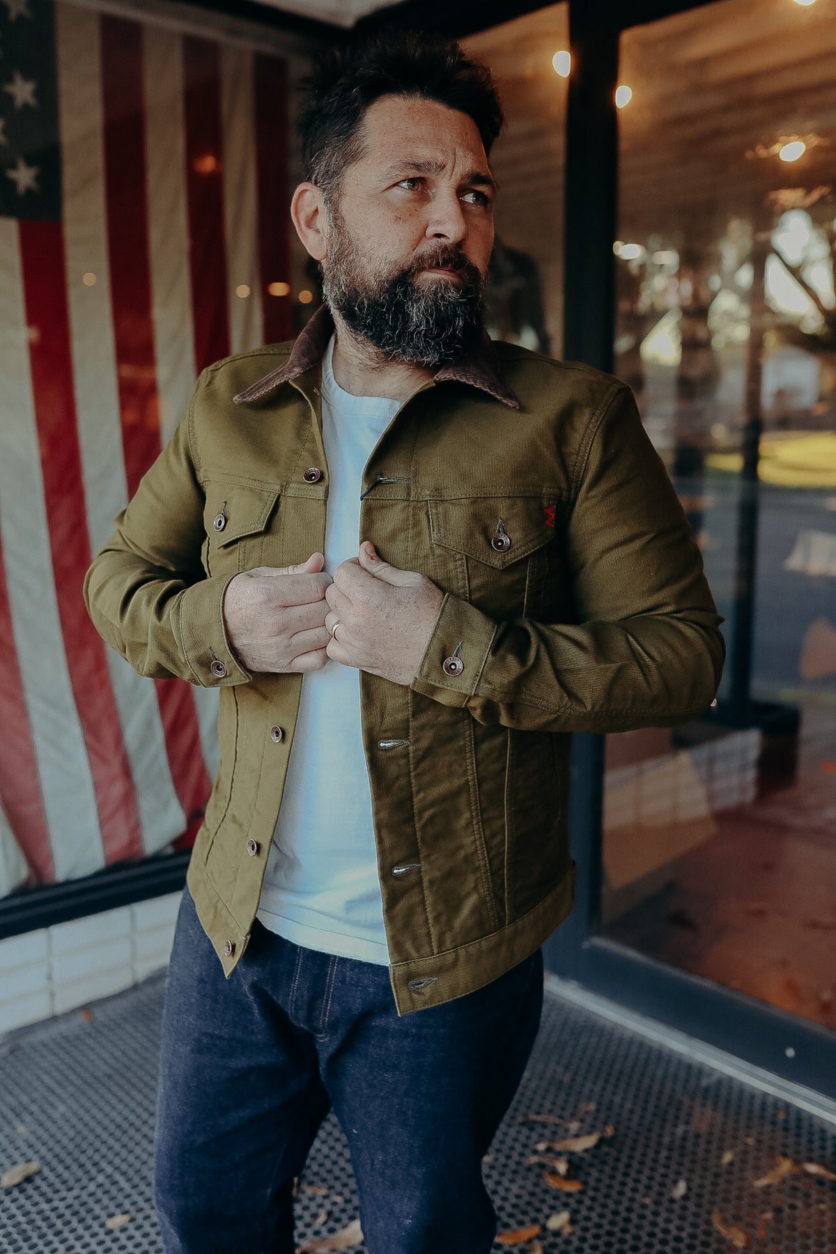Bearded man wearing olive green work jacket and white t-shirt standing by American flag in casual outdoor setting