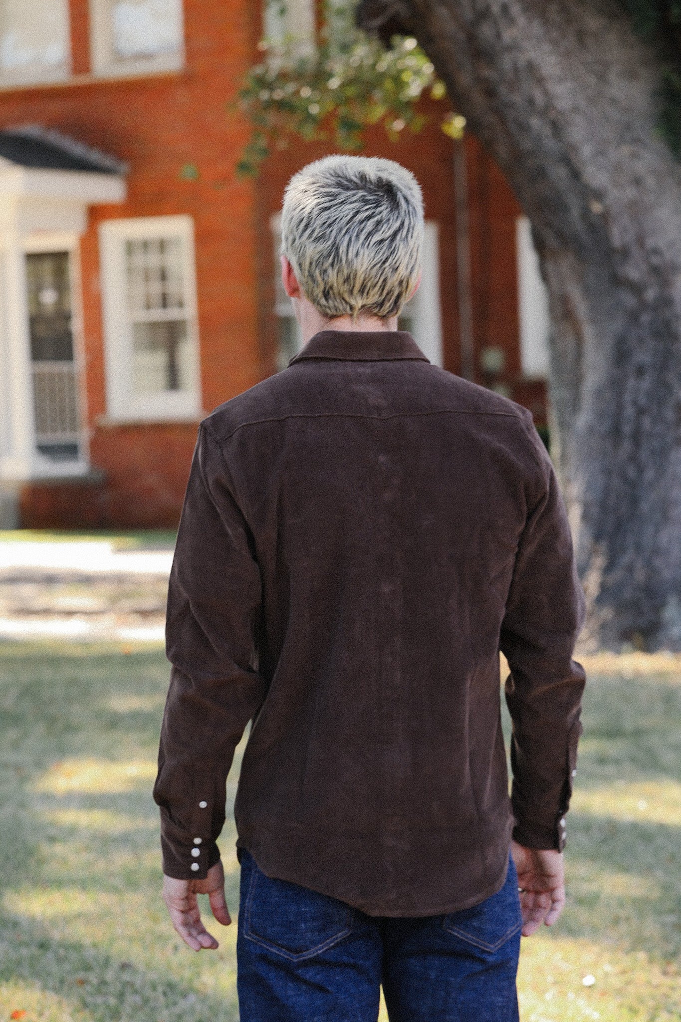 Back view of a person with short bleached hair wearing a brown suede shirt and blue jeans outdoors