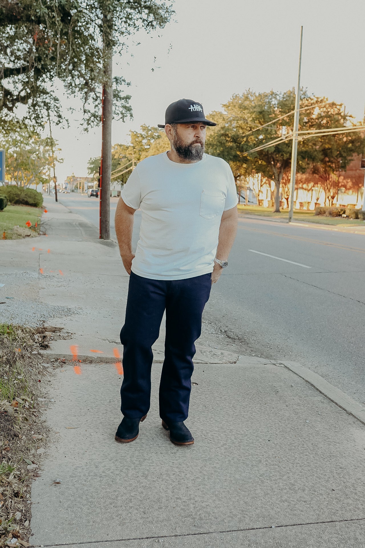 Man with beard wearing white t-shirt, navy pants, black cap, and boots standing on sidewalk near road