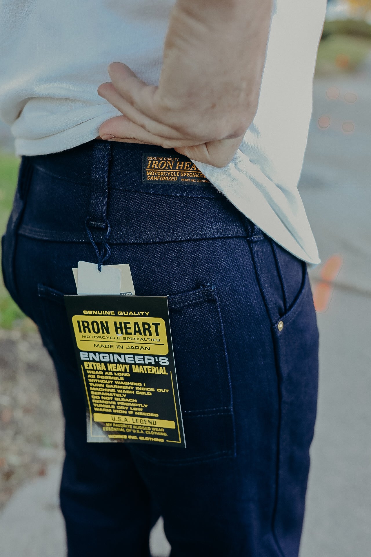 Close-up of person wearing dark blue Iron Heart jeans with yellow and black branded tag hanging from back pocket