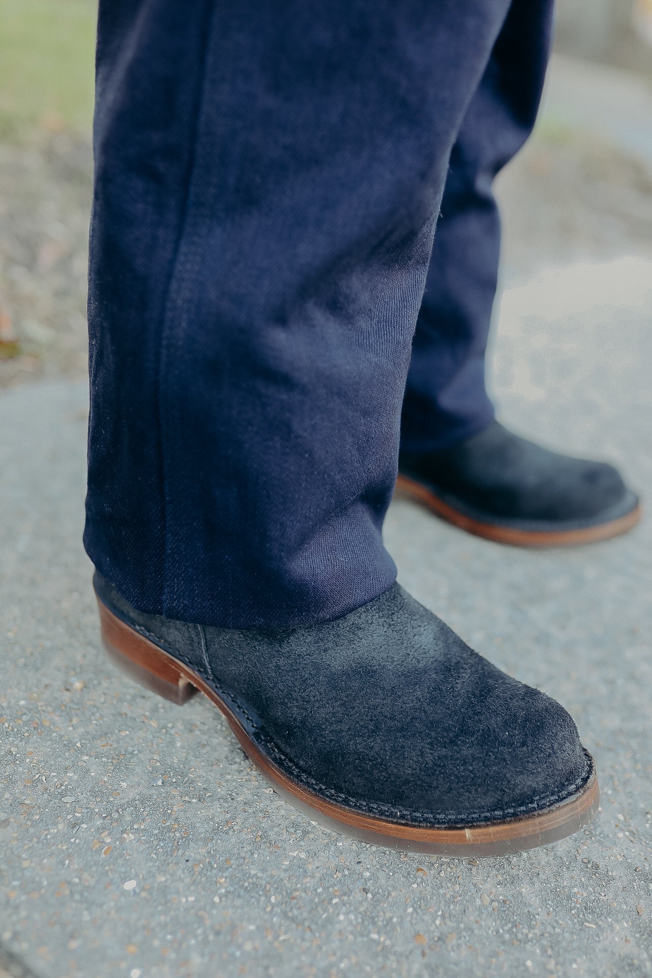 Close-up of black suede leather boots with brown sole worn with dark blue pants on concrete surface