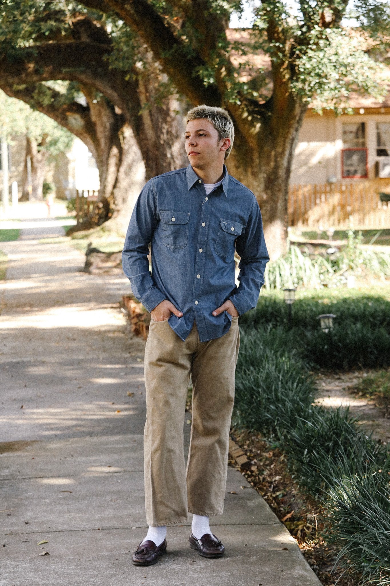 Man wearing blue denim shirt, khaki pants, and loafers walking on tree-lined sidewalk in daylight