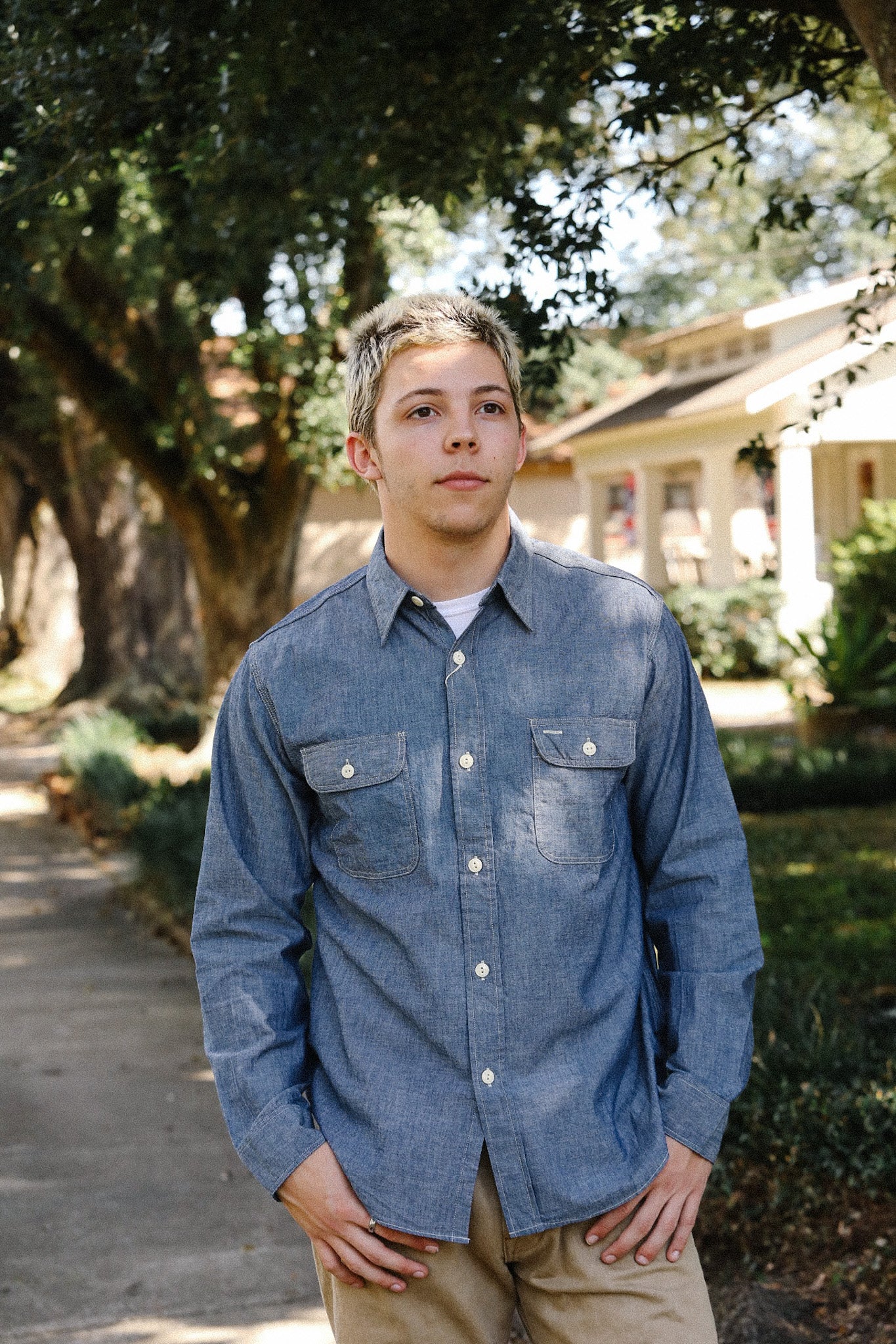 Young man wearing a blue chambray button-up shirt and khaki pants standing outdoors near trees and house