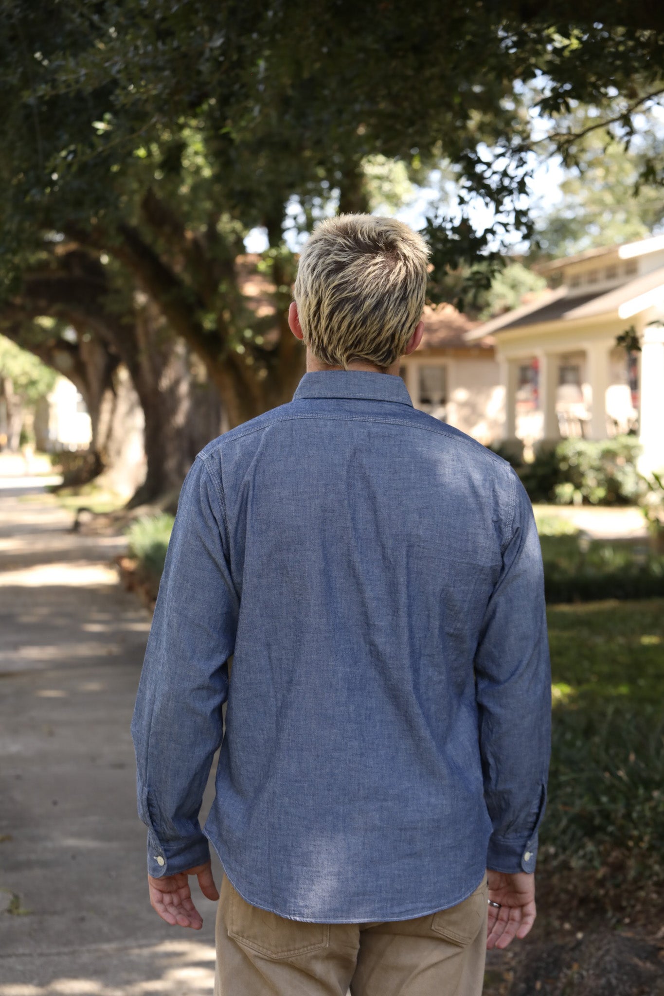 Man with dyed blond hair wearing a blue chambray shirt and tan pants walking on a tree-lined suburban sidewalk