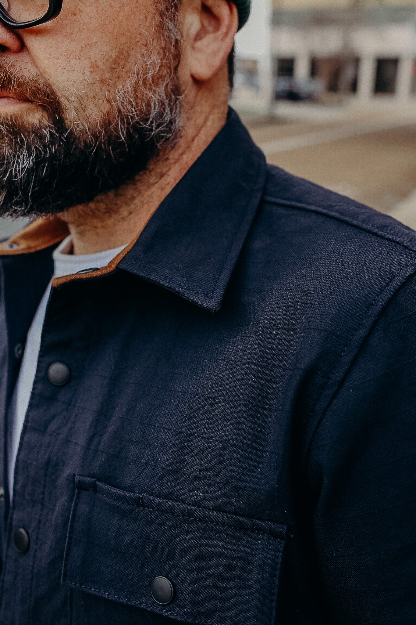Close-up of a man with a gray beard wearing a dark navy jacket with snap buttons and a brown collar detail