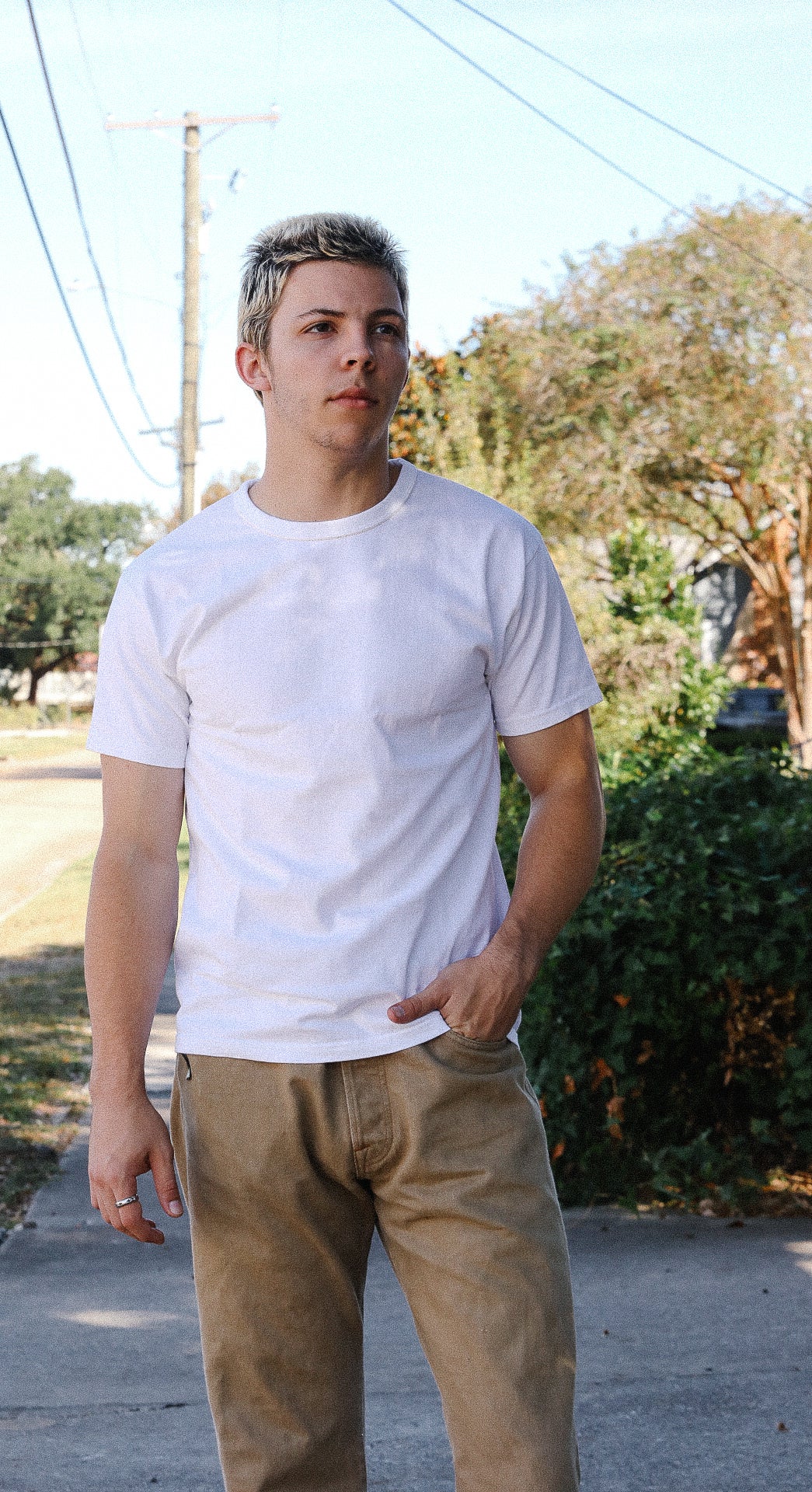 Young man wearing plain white t-shirt and tan pants standing outdoors on sidewalk with trees and power lines