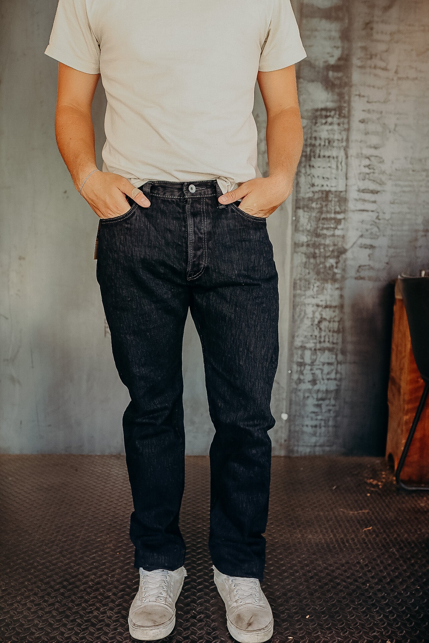 Person wearing dark raw denim jeans with hands in pockets, white sneakers, and a beige t-shirt in an industrial setting