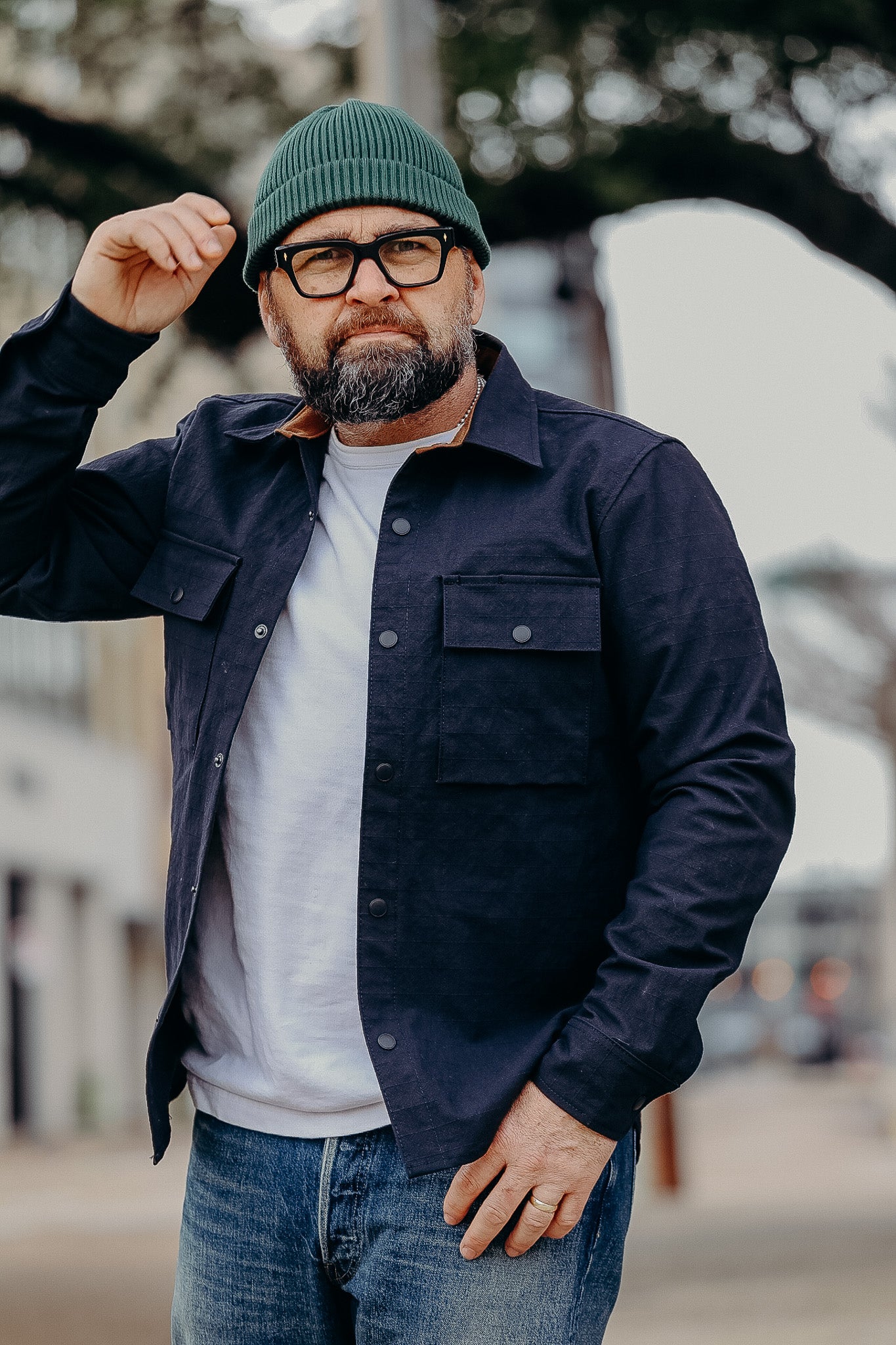 Man wearing green beanie, black glasses, navy jacket over white shirt, and blue jeans in urban setting