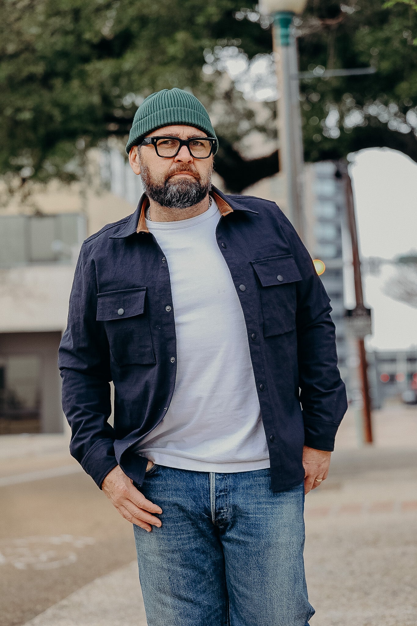 Man wearing green beanie, glasses, navy shirt jacket over white t-shirt, and jeans outdoors on urban sidewalk