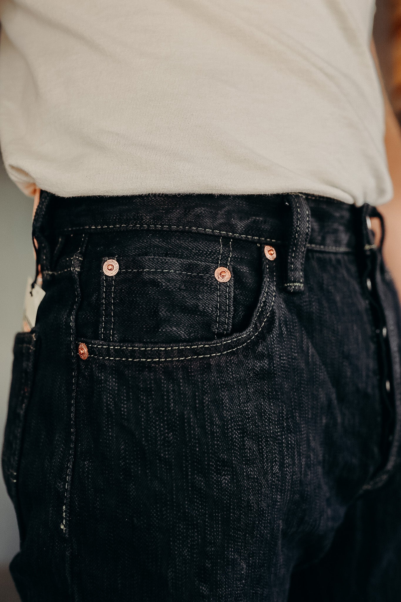 Close-up of black denim jeans with copper rivets and detailed stitching worn with a light beige shirt
