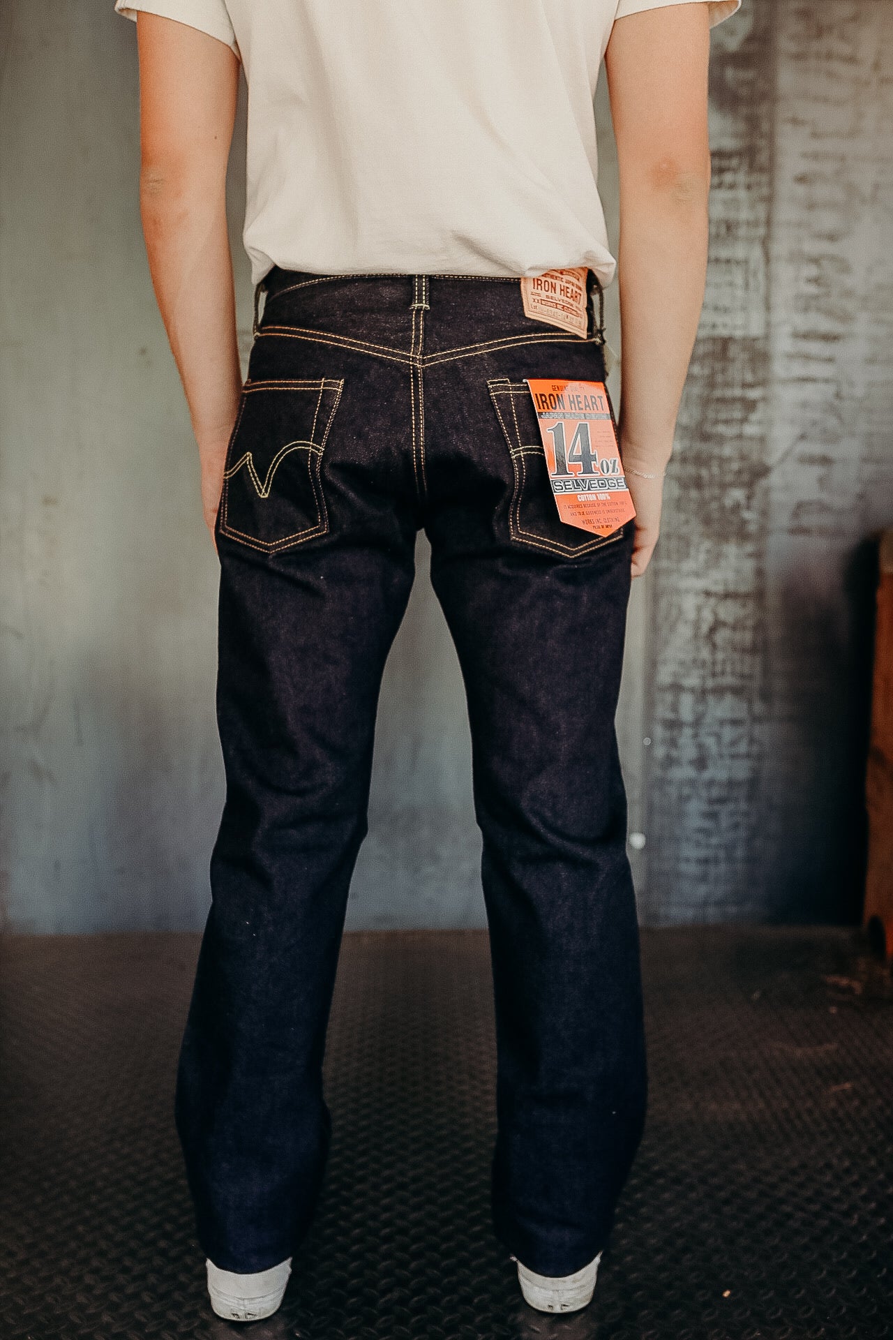 Back view of person wearing Iron Heart 14oz selvedge denim jeans with tags in industrial setting